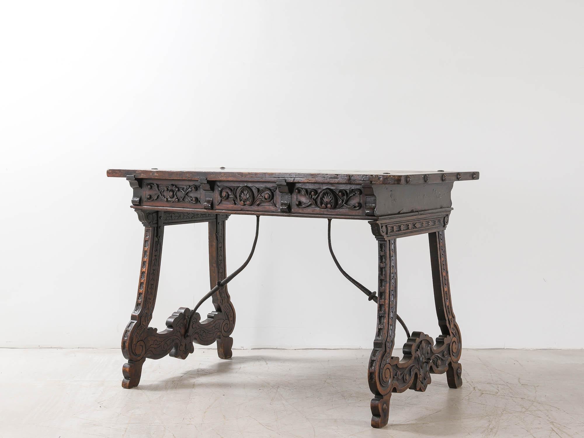 An ornately carved Spanish 19th century Neo-Renaissance table in dark chestnut. With a beautifully patinated top with iron rivets securing it to a three drawer frieze with floral carving and velvet backed iron drawer pulls. Sitting on carved trestle