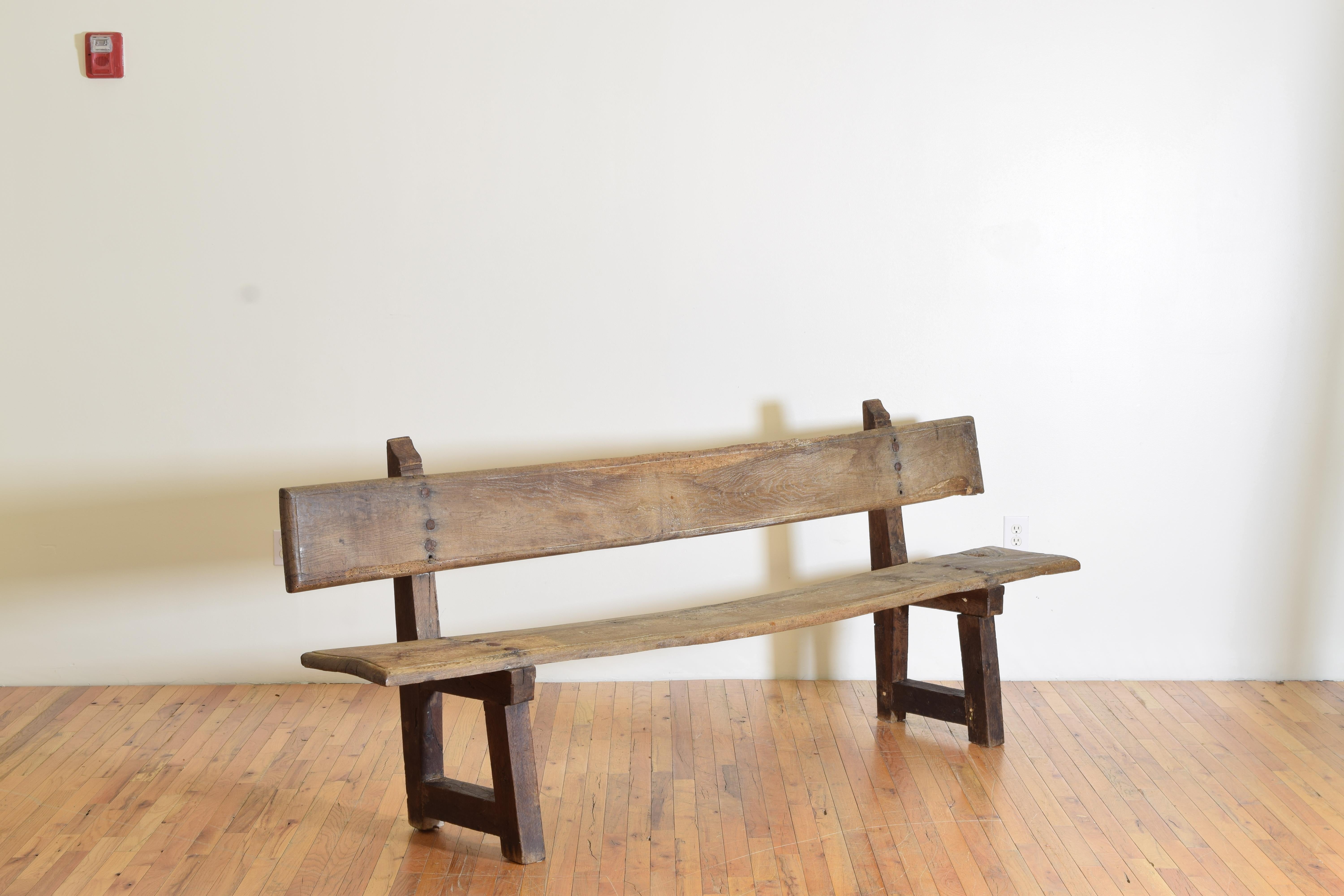 In a primitive style this bench was originally built for a hallway or perhaps even a private chapel, having two trestle form supports with stylized finial tops supporting plank seats and backs, the planks having molded edges and joined to the bases