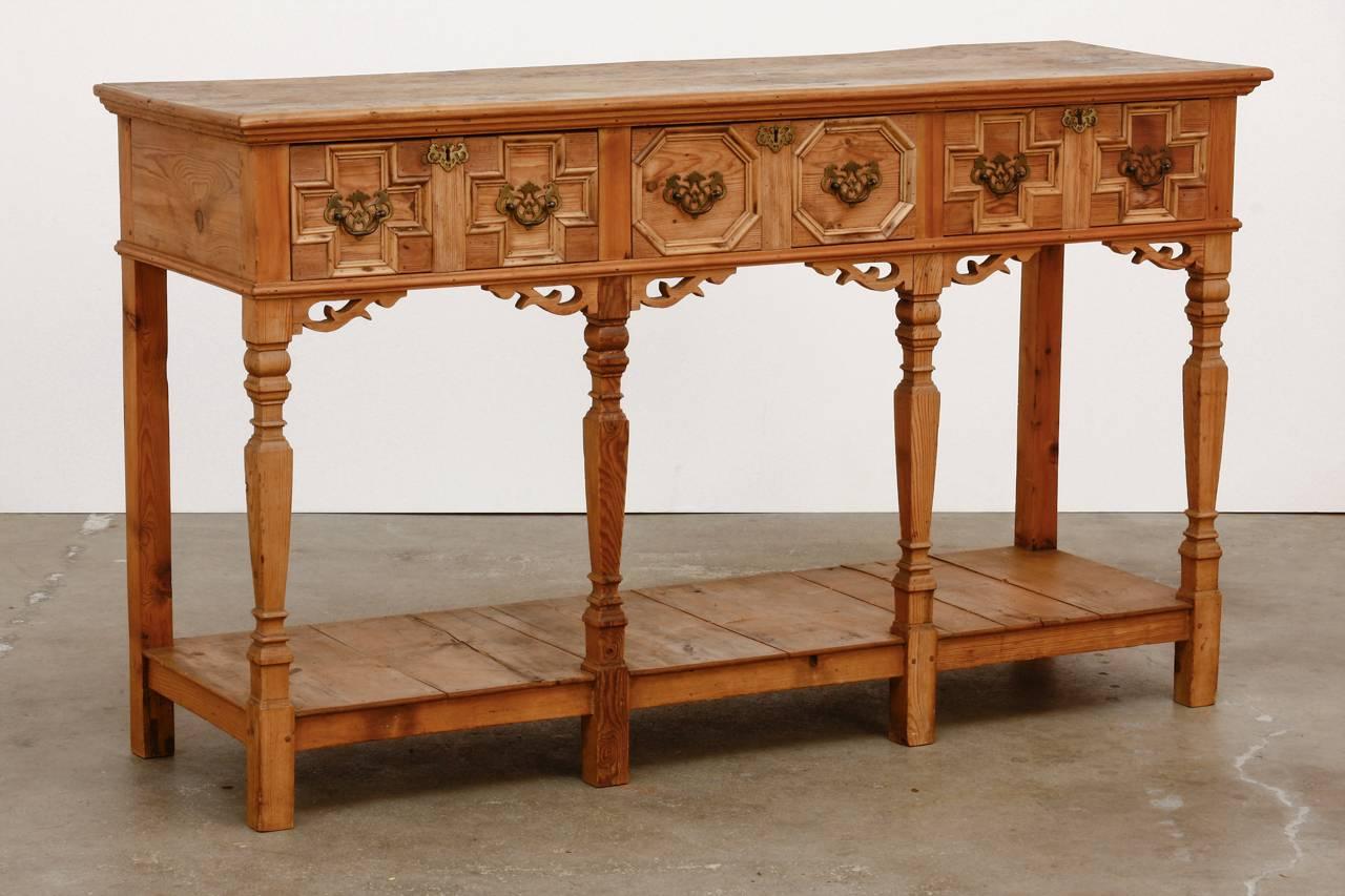 Rustic Spanish Baroque style console table or server constructed  from pine. Features a three drawer front decorated with geometrical designs and brass pulls. Supported by six square legs the front being tapered and carved. There is a pot rack on