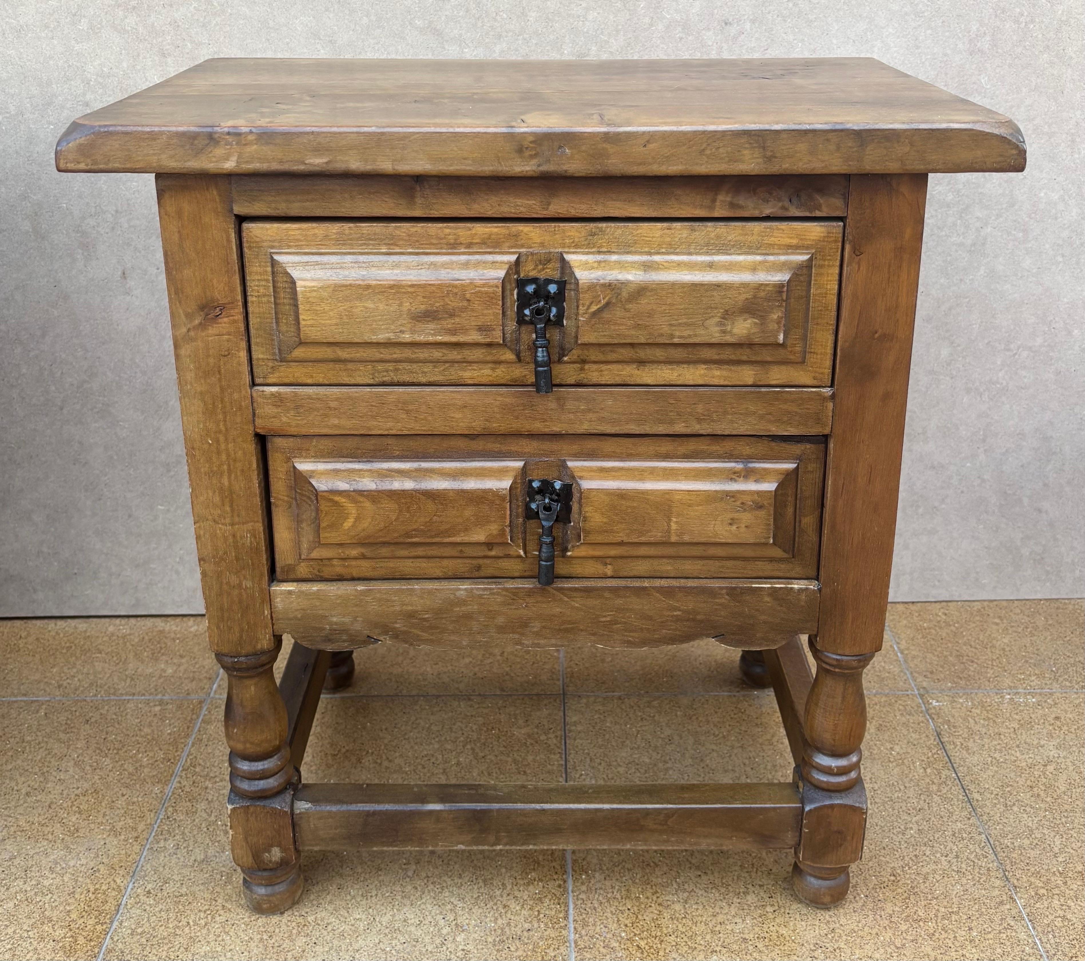 Paire de tables de nuit rustiques espagnoles du milieu du 20e siècle, fabriquées en pin massif avec une finition chaude dans les tons de noyer. Chaque pièce comporte deux façades de tiroir moulées, agrémentées de ferrures traditionnelles en fer