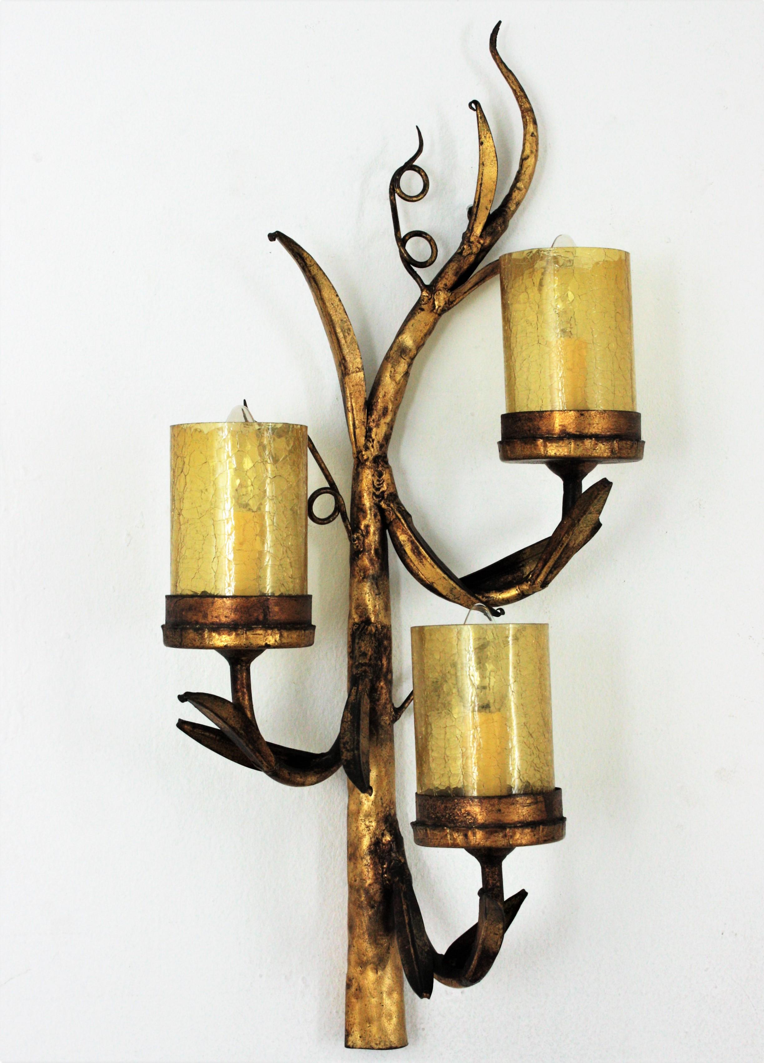 Three-arm branch wall light / foliage wall sconce with amber cracked glass cylinder shades, Spain, 1950s.
This brutalist wall sconce features a tree form light fixture with three branches holding cylinder glass shades. Foliage details adorning the