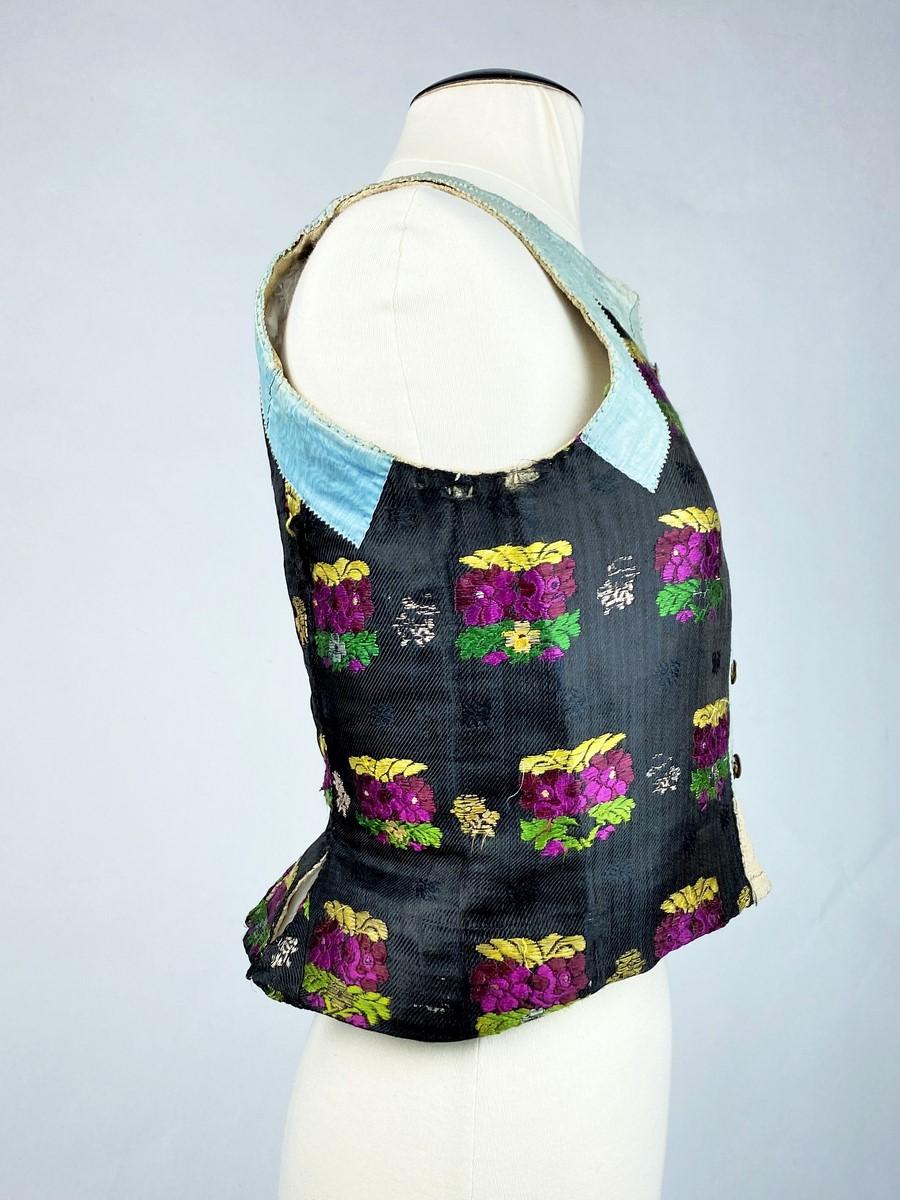 First half of the 19th century
Spain

Precious caraco or bodice in silk twill embroidered with bouquets, fully lined with unbleached hemp. Sleeveless caraco buttoned at the front and highlighted with applied ribbons in rainbow-blue moiré silk