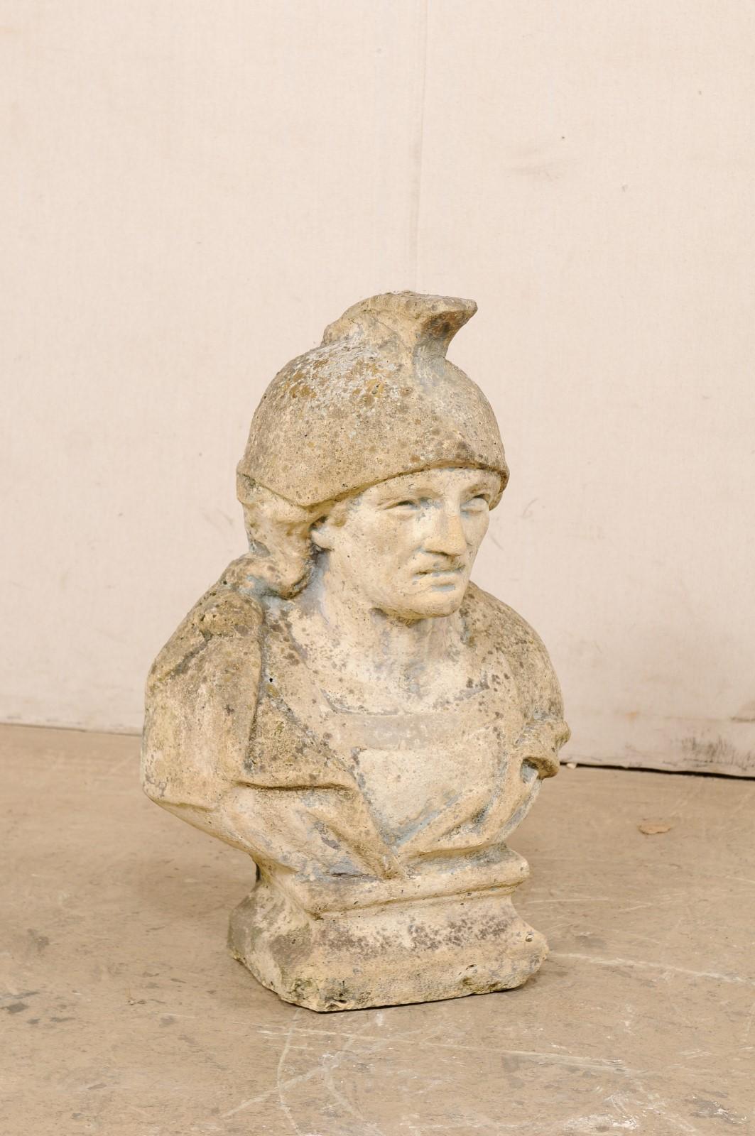 A Spanish cast-stone bust art piece. This vintage decoration from Spain, created from cast stone, features a male bust, in military attire with Roman-style helmet. There is a nicely weathered patina, and small losses throughout this piece, giving it