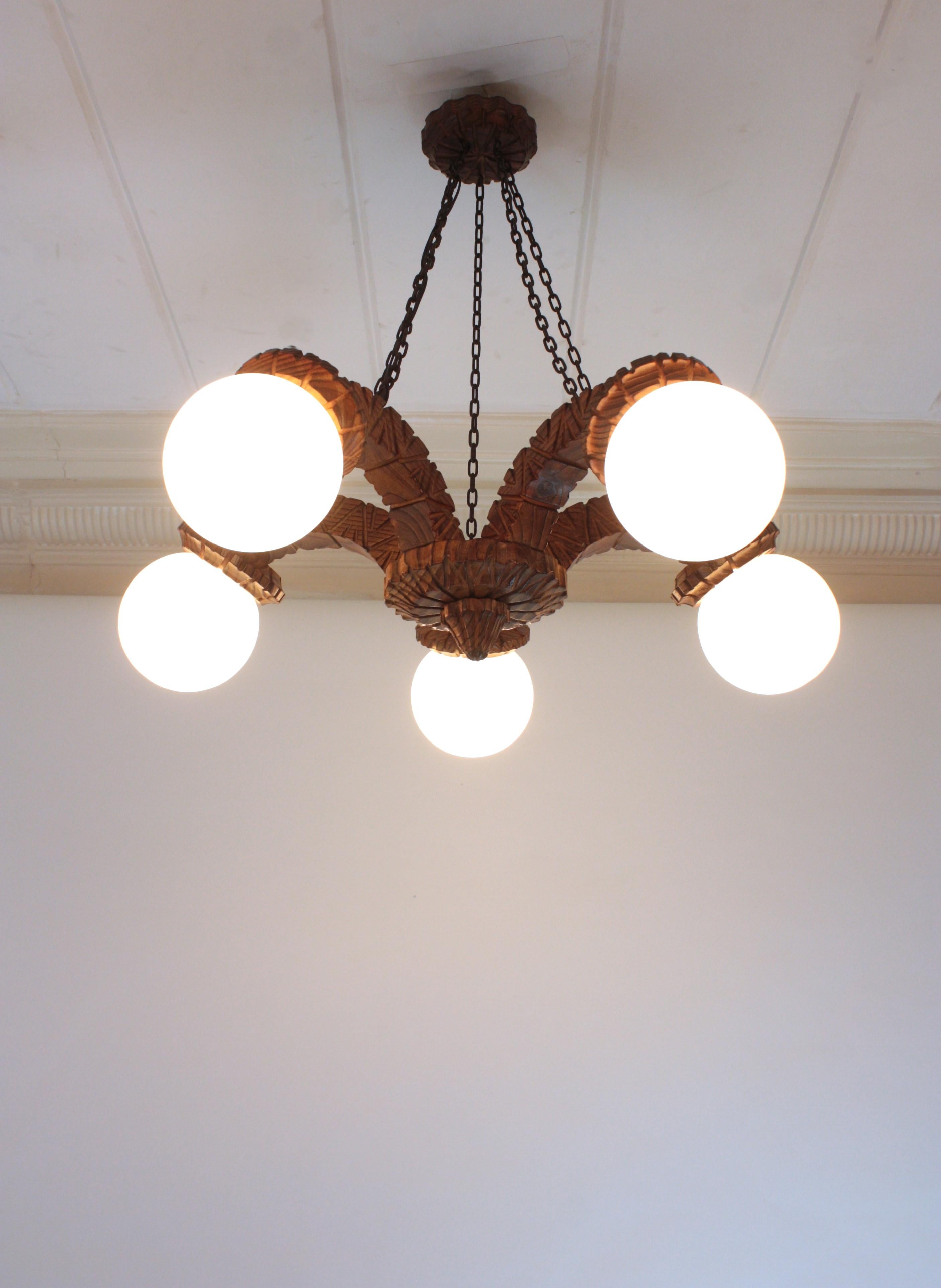 Lampadario Coloniale Spagnolo in vetro opalino a cinque bracci, Wood intagliato
Spagna, 1940-1950.
Dimensione grande.
Lampadario in legno di pino intagliato a mano con 5 paralumi a globo in vetro di latte. 
Questo splendido lampadario / plafoniera a