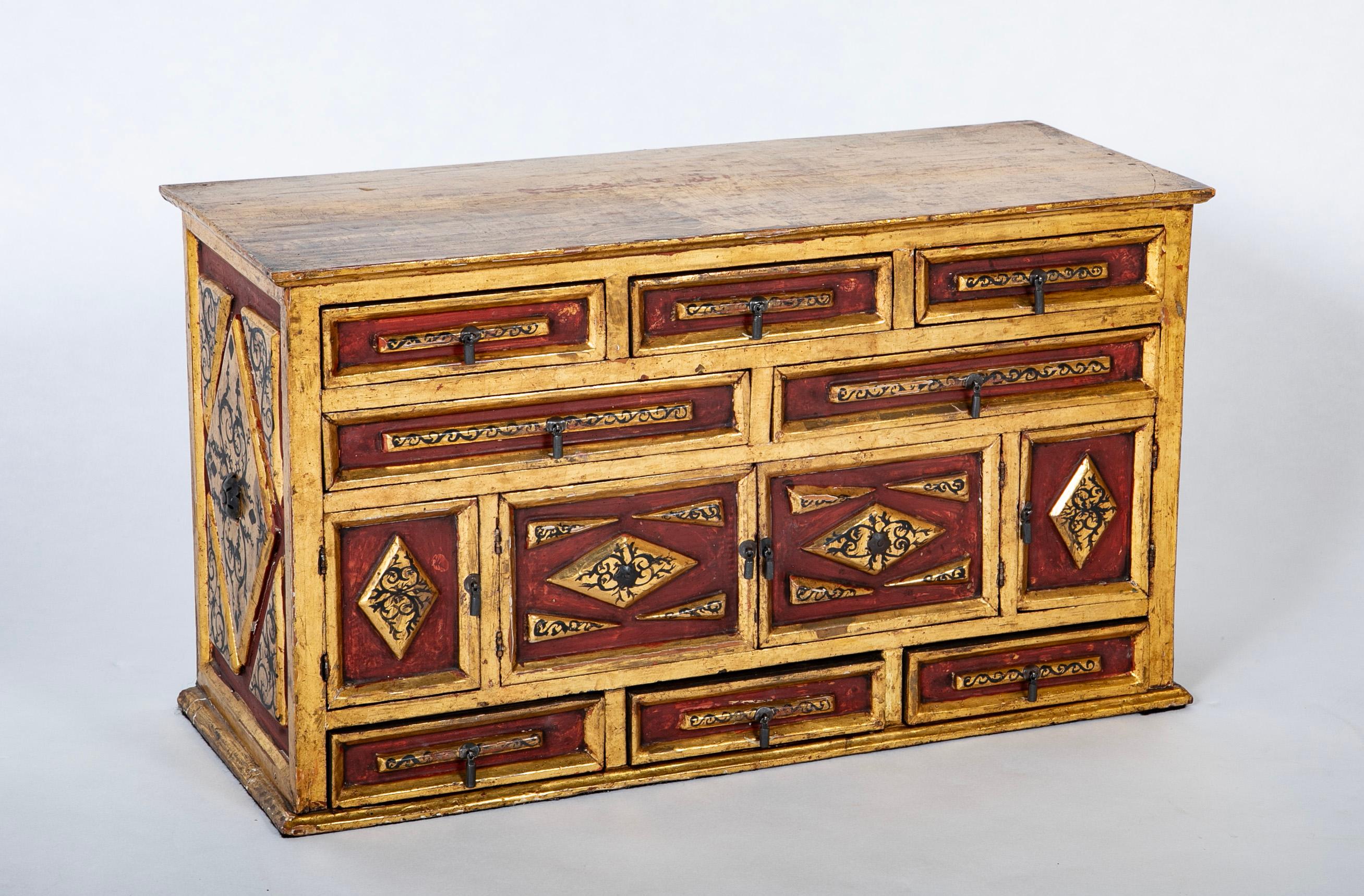 Tablero vargueno de madera pintada y dorada de estilo colonial español, arcón con ocho cajones y cuatro puertas con tiradores de gota de hierro. Un cofrecito dulce y con mucha personalidad, decorado con pintura roja y diseños arabescos negros.