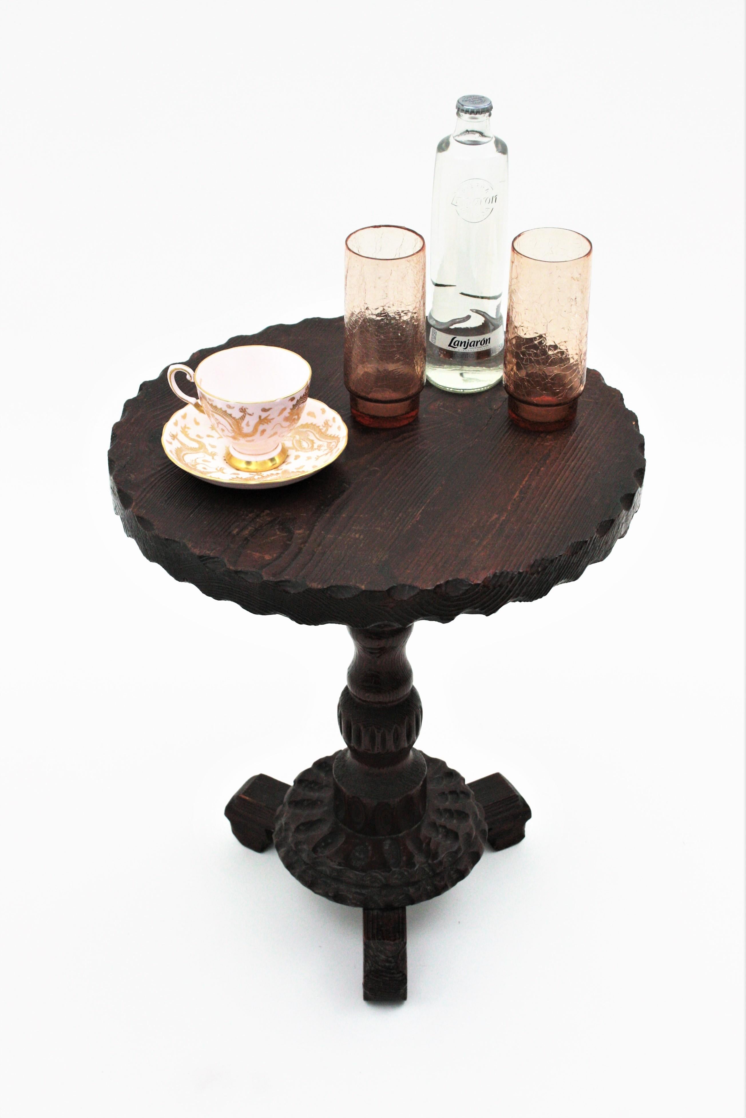 Hand-carved pine wood gueridon table or occassional table standing on tripod base. Spain, 1940s.
The tabletop in round shape has scalloped details on the edge. It is supported on a turned wood stem and three carved legs.
This table has a rustic