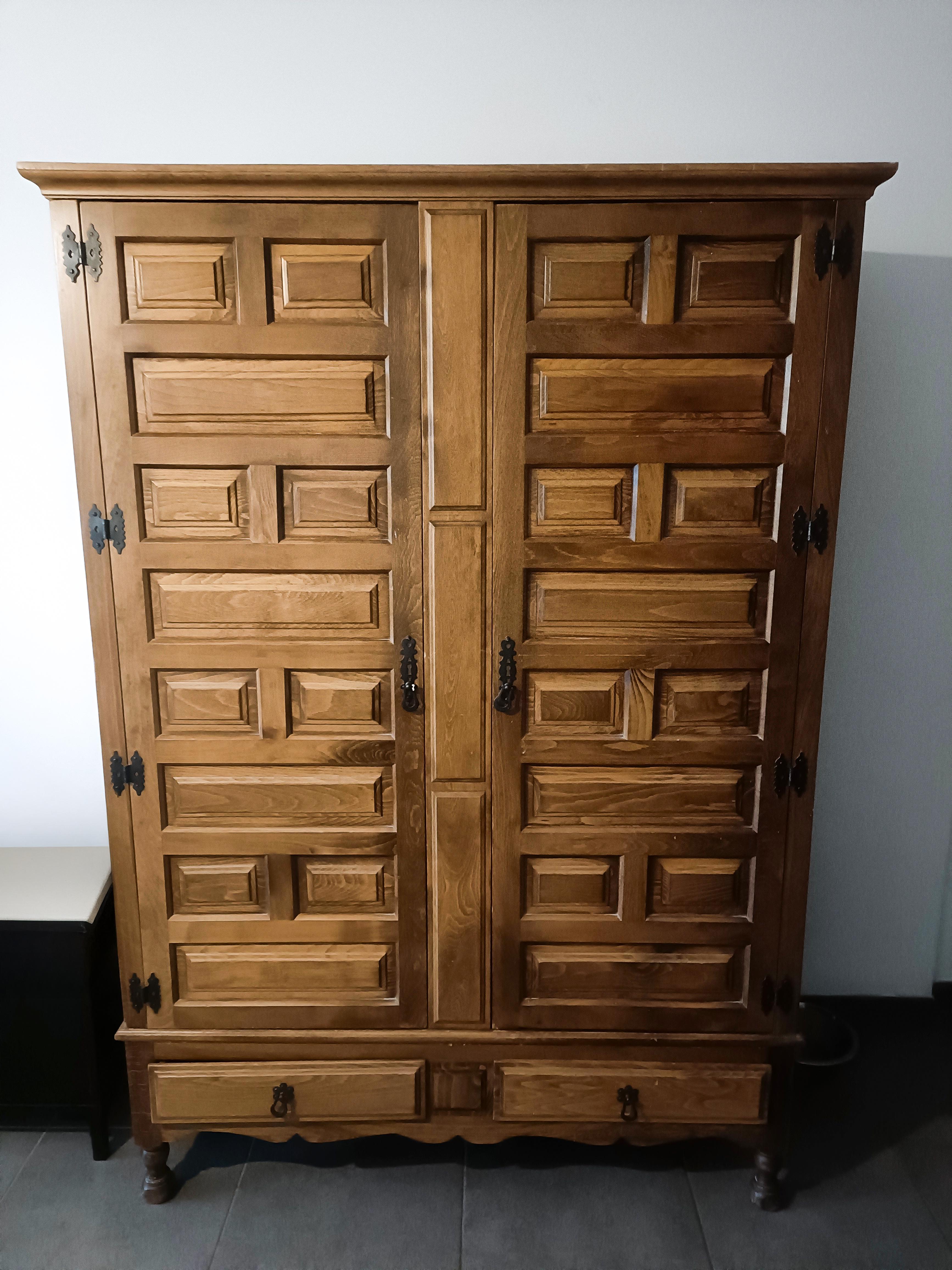 This striking wardrobe showcases the influence of Spanish Colonial Revival design, marked by its bold geometric paneling, intricate woodwork, and dark wrought iron hardware. The doors feature deeply set rectangular carvings arranged in a rhythmic,
