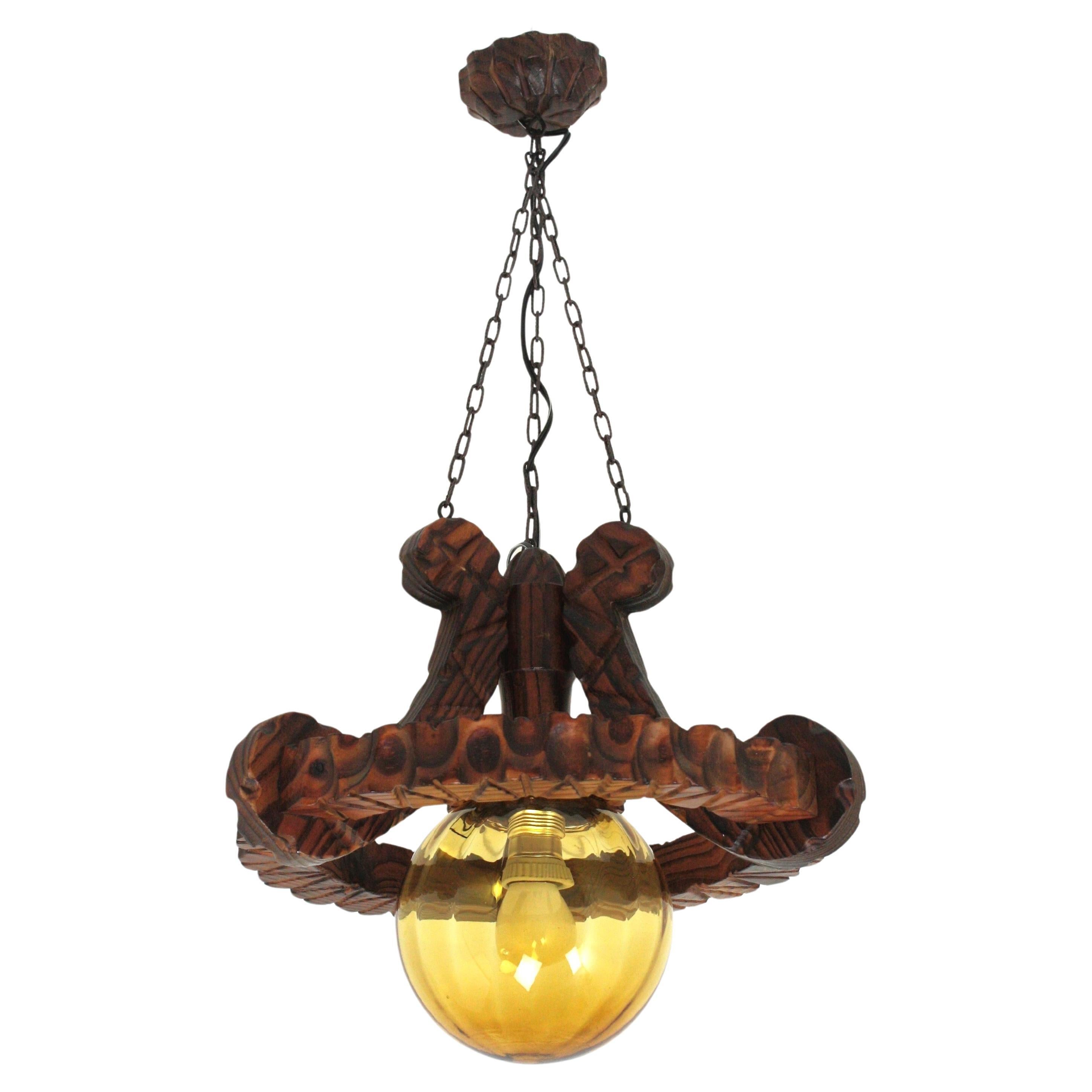 Handcarved pine wood ring ceiling light fixture with amber glass globe lampshade. Spain, 1940-1950.
This ceiling light pendant lamp has an eye-catching construction. The ring backplate in carved wood holds an amber glass globe shaped lampshade at