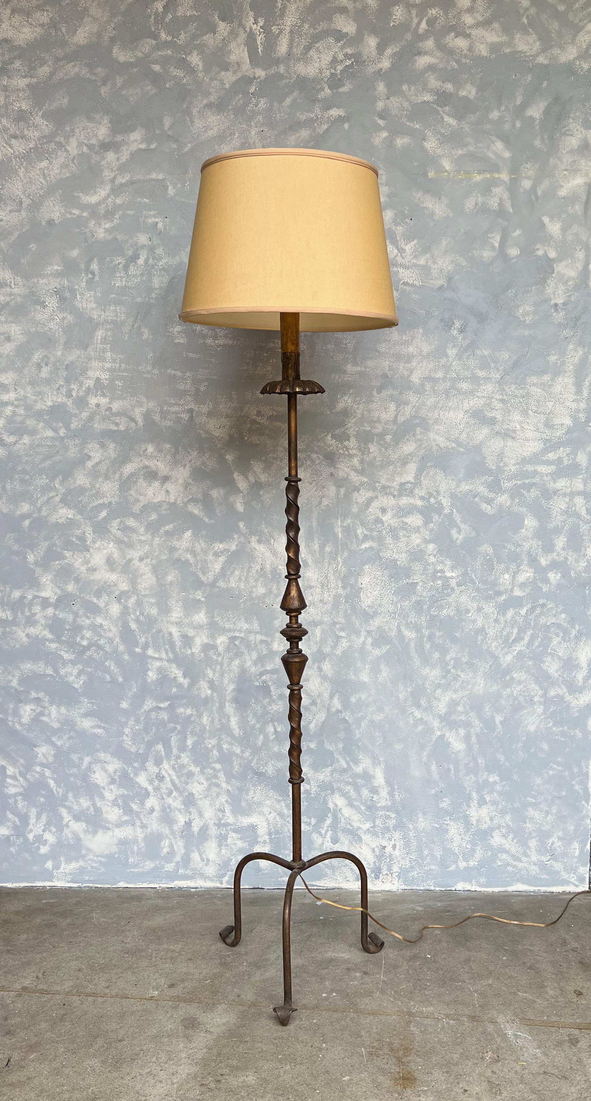 This Spanish gilt iron floor lamp, dating back to the 1950s, is a distinctive piece with its elevated tripod base. The lamp possesses an aged darkened gold patina that adds character and an air of antiquity. The stem's upper part features a double