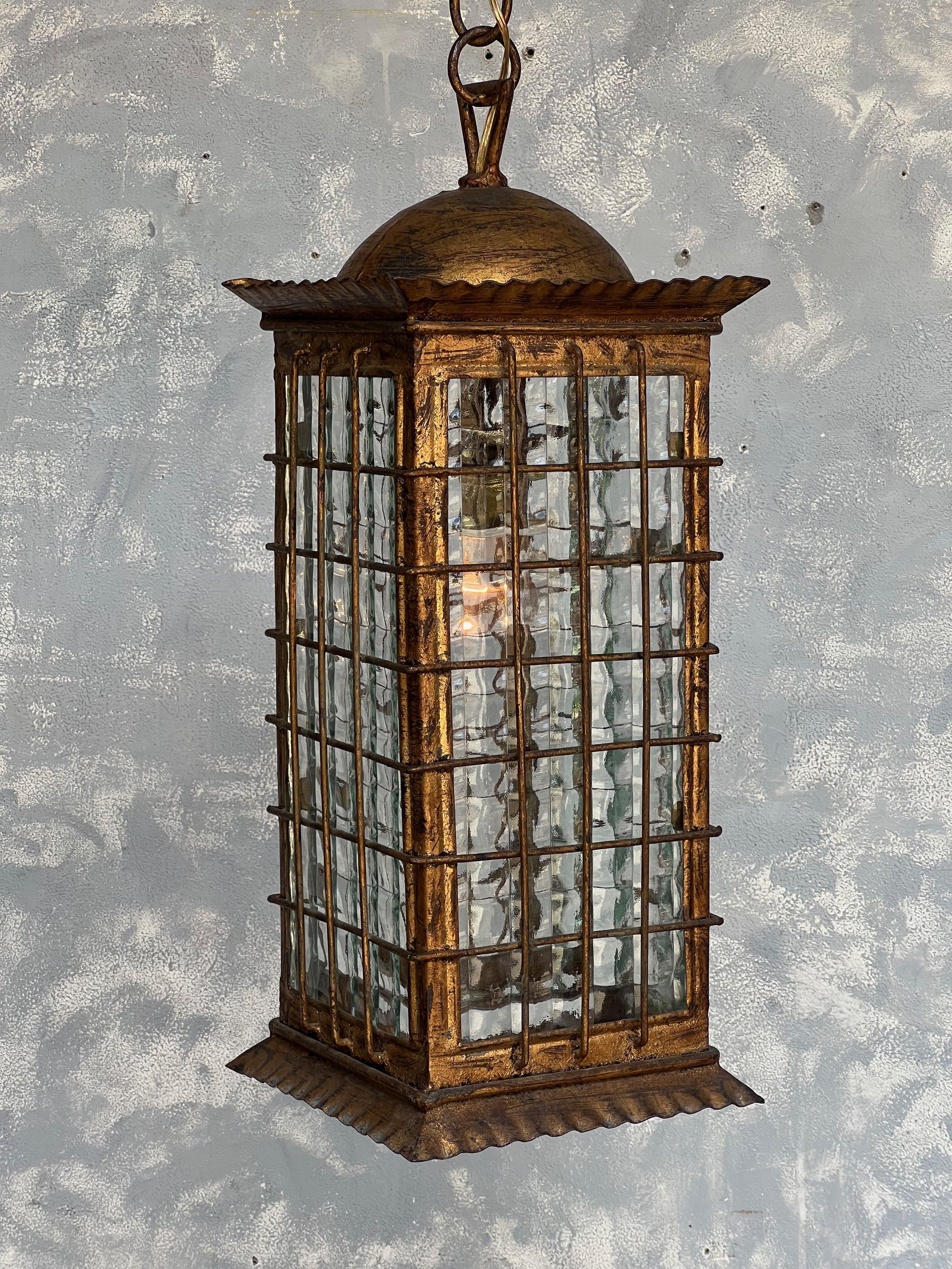 A stunning Spanish gilt metal lantern from the 1960s, boasting exquisite craftsmanship and timeless appeal. This lantern features textured glass, creating a beautiful reflective effect when illuminated. It is elegantly suspended by a chain, which is