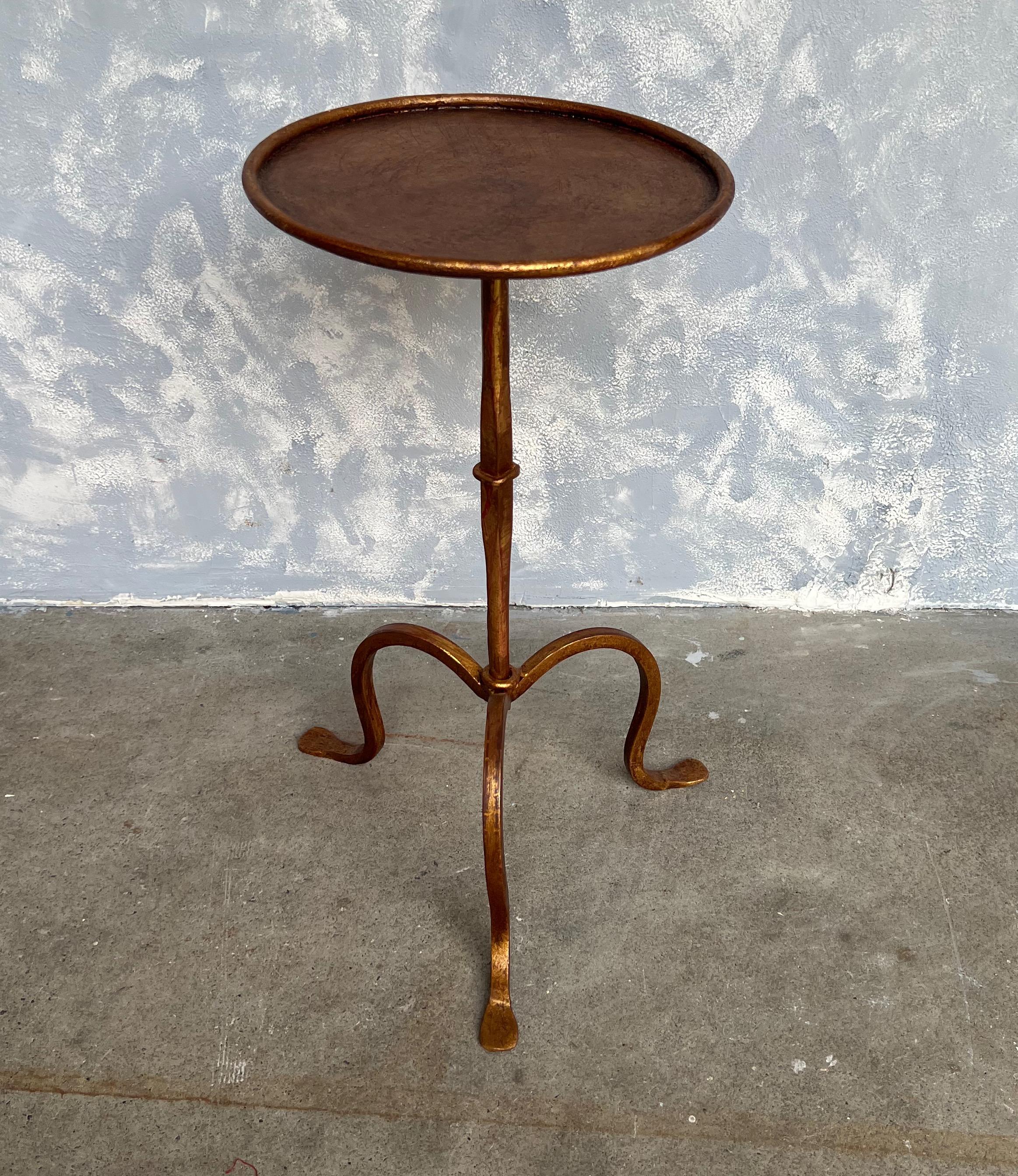 This Spanish 1950s gilt iron drinks table features a unique inverted tapered stem, intersected in the middle by a circular design element. The table sits on an elegant tripod base, and its original finish is a hand-applied gold patina with red