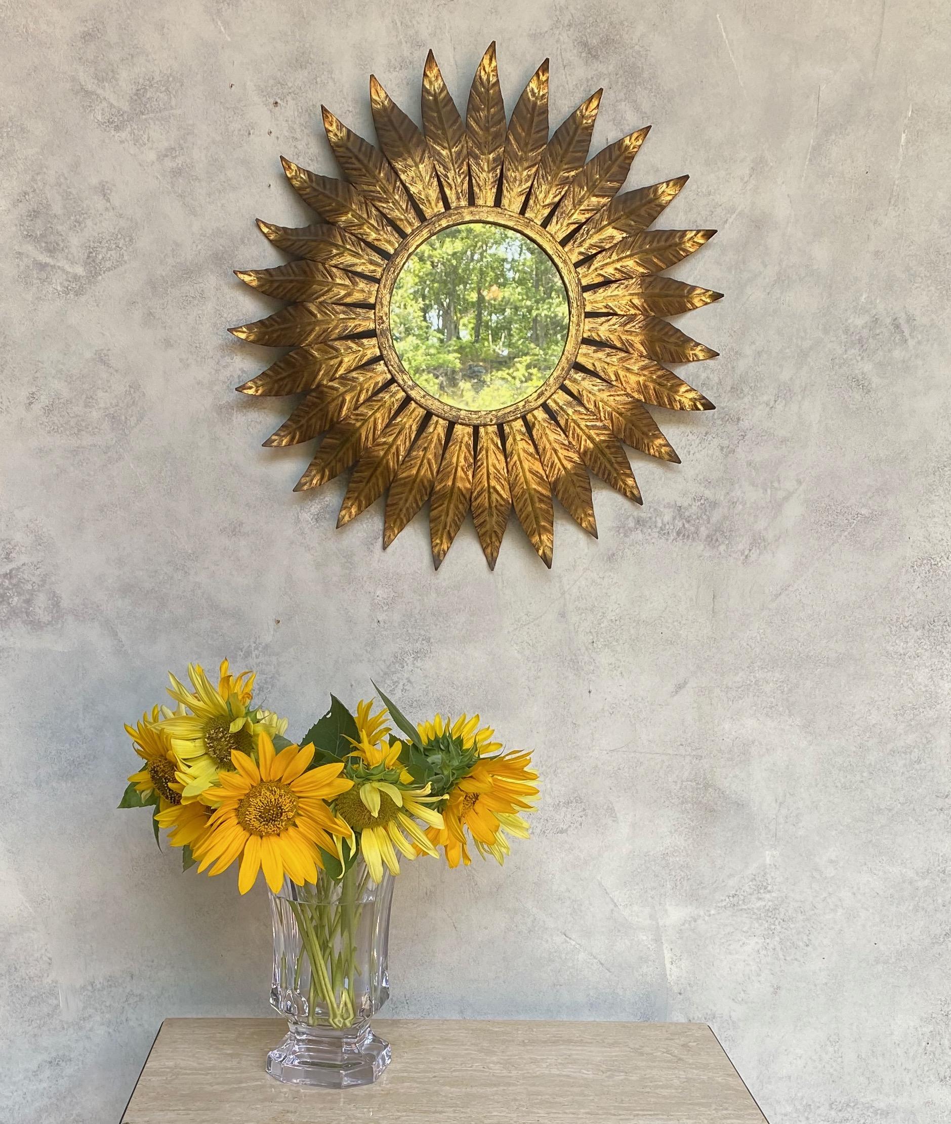This round gilt metal sunburst mirror, originating from Spain in the 1950s, features long narrow leaves converging in the middle and radiating from a solid border, creating a stunning visual effect. The original glass, showing signs of age, adds to