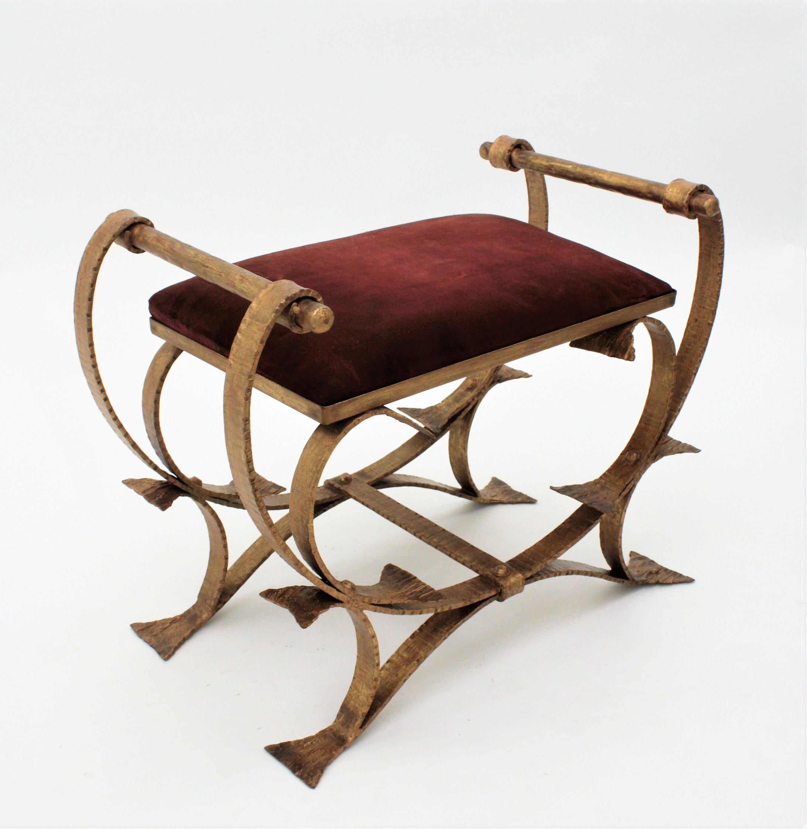 Sculptural hand forged iron stool with arms upholstered in burgundy red velvet, Spain, 1920s-1930s.
This gorgeous two handled bench is beautifully constructed in hand forged iron finished in gold leaf gilding.
Wearing its original burgundy velvet