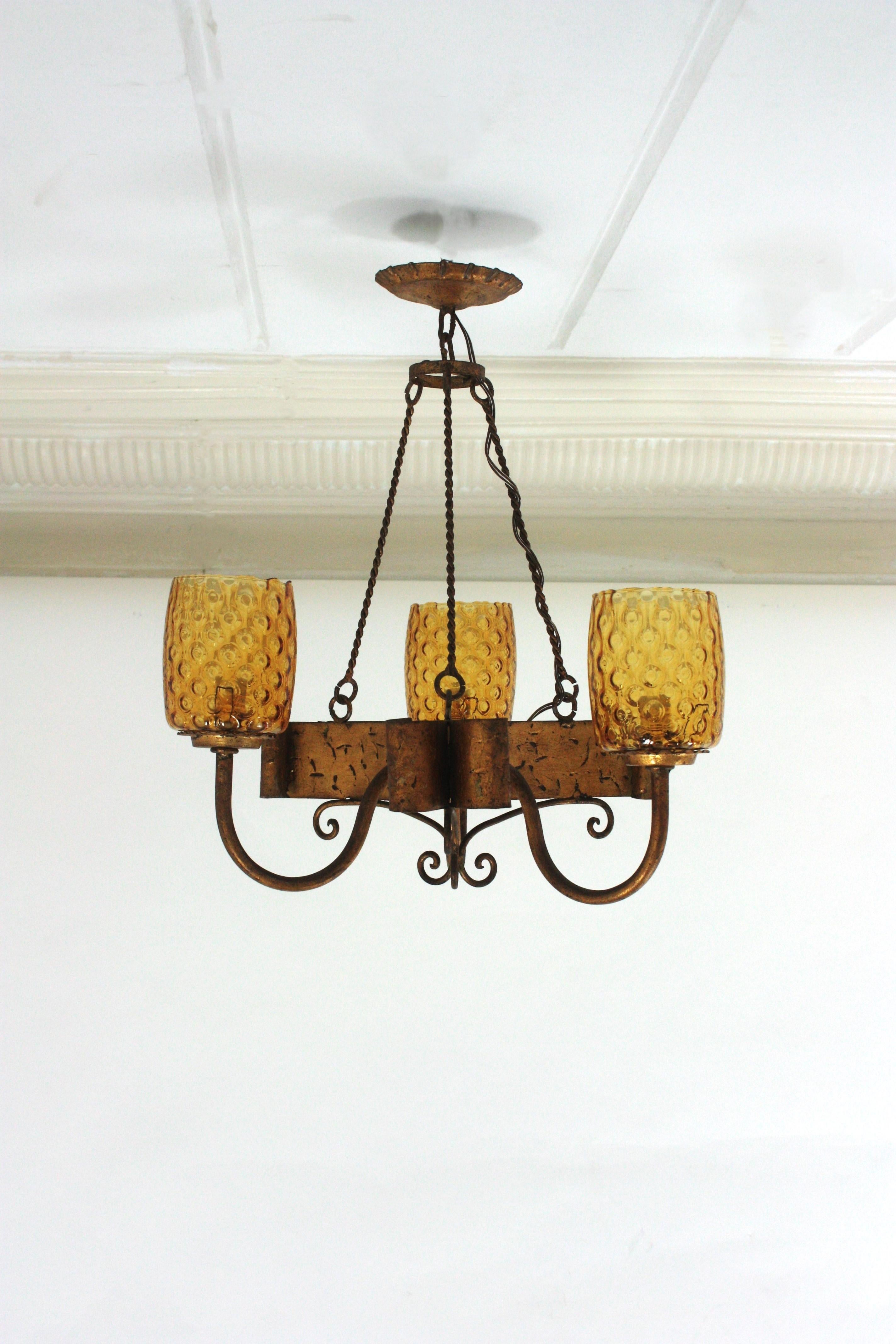 Hand Forged Gilt Iron and Amber Glass Spanish Chandelier
Hand forged iron three-light pendant or ceiling hanging light with amber lampshades, Spain, 1930s-1940s.
This eye-catching chandelier features a wrought iron structure with three arms holding