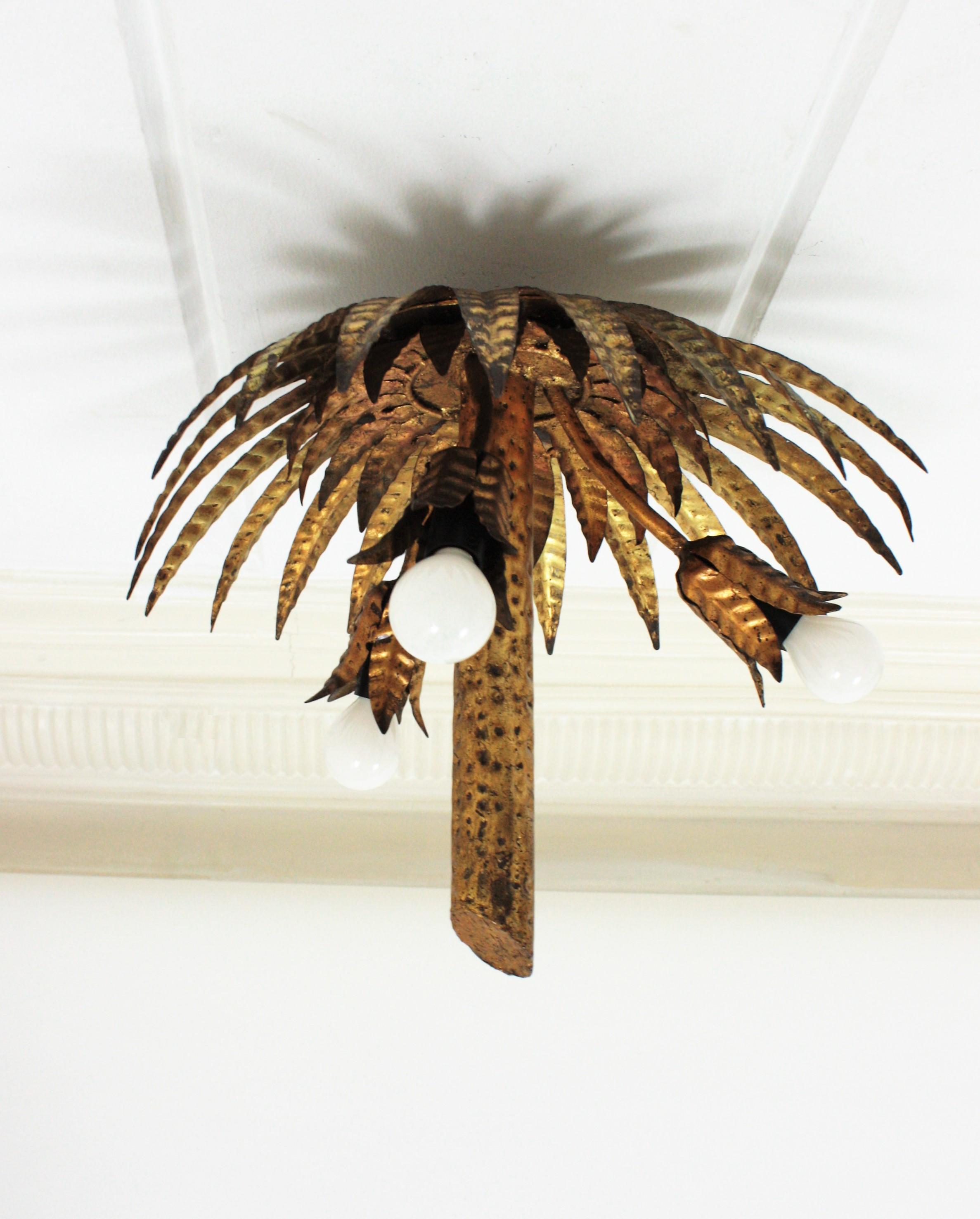 Eye-catching hand-hammered gilt iron palm tree ceiling light fixture or pendant. Spain, 1950s.
This amazing piece has two layers of leaves and 3 branches finishing with flowers as bulb holders. The lamp wears its original vintage patina with gold