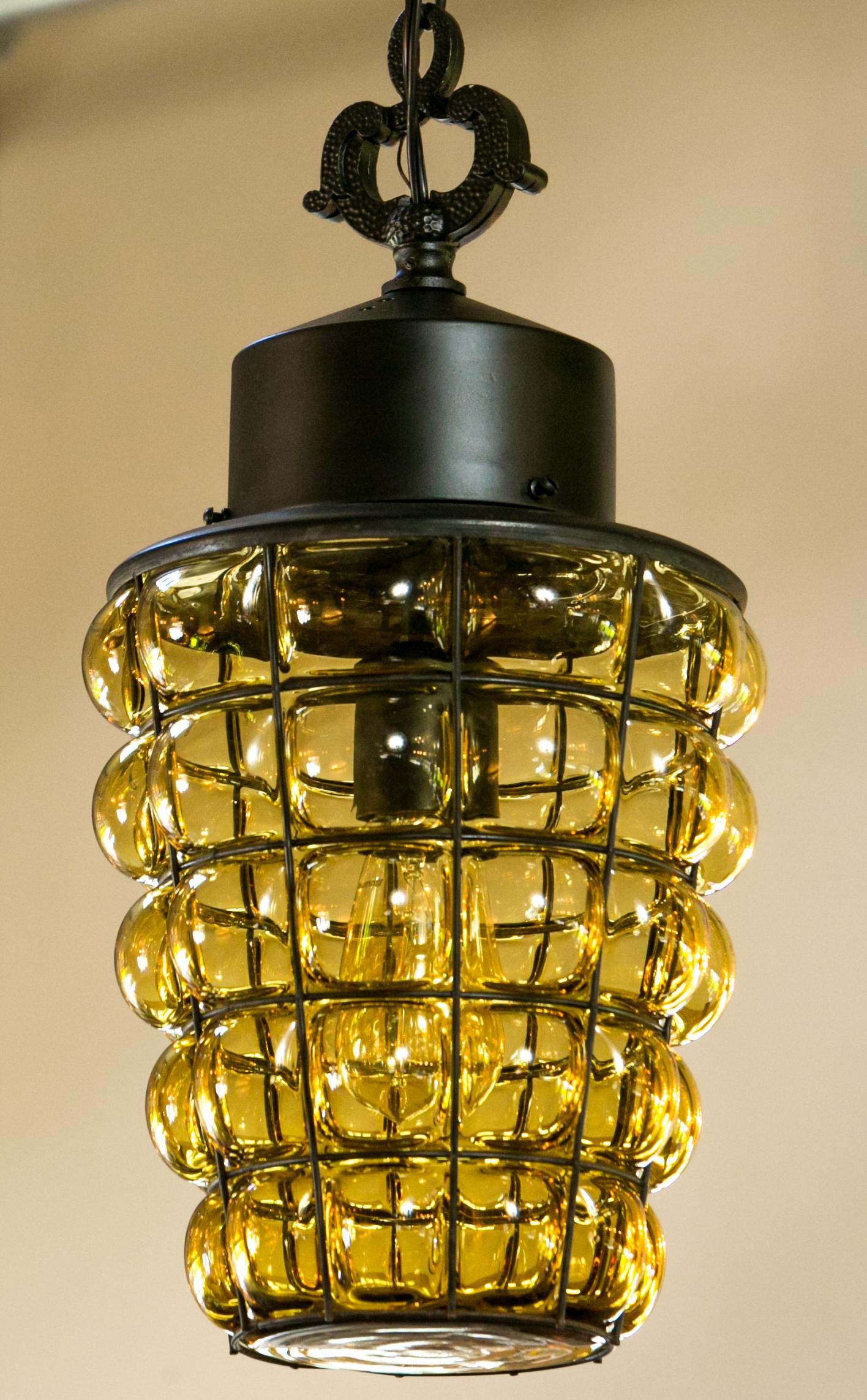 Spanish Colonial caged glass lantern with single medium-base (Edison) light. Black painted wrought iron with a yellow/amber transparent glass piece encased by a wrought iron honeycomb style frame. In this style of light the glass is generally hand