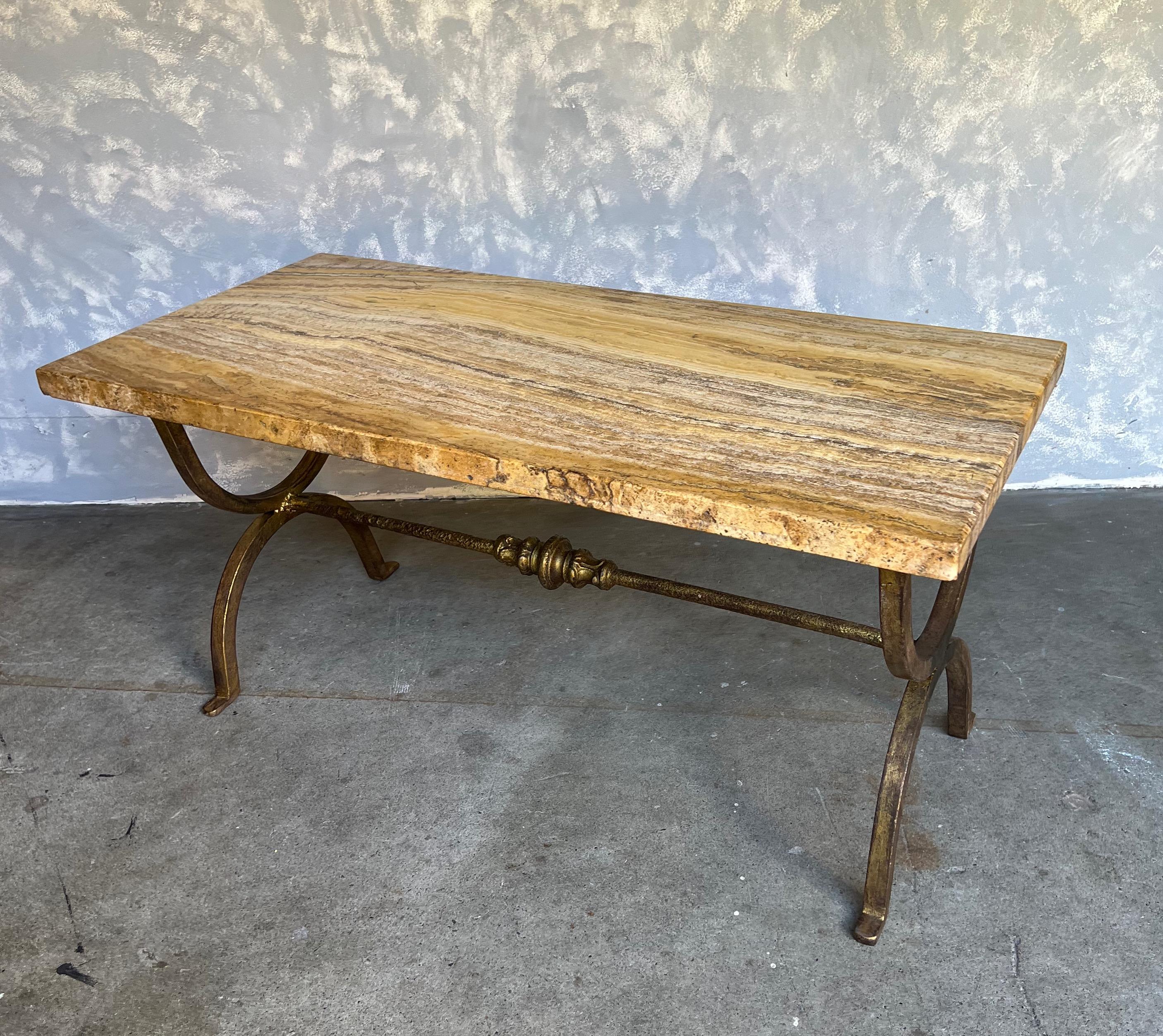 A heavy iron coffee table with a thick marble top. The gilt forge iron base has a central stretcher with a classical decorative center motif. The marble top is a richly veined golden color with brown and yellows patterns. Very good vintage