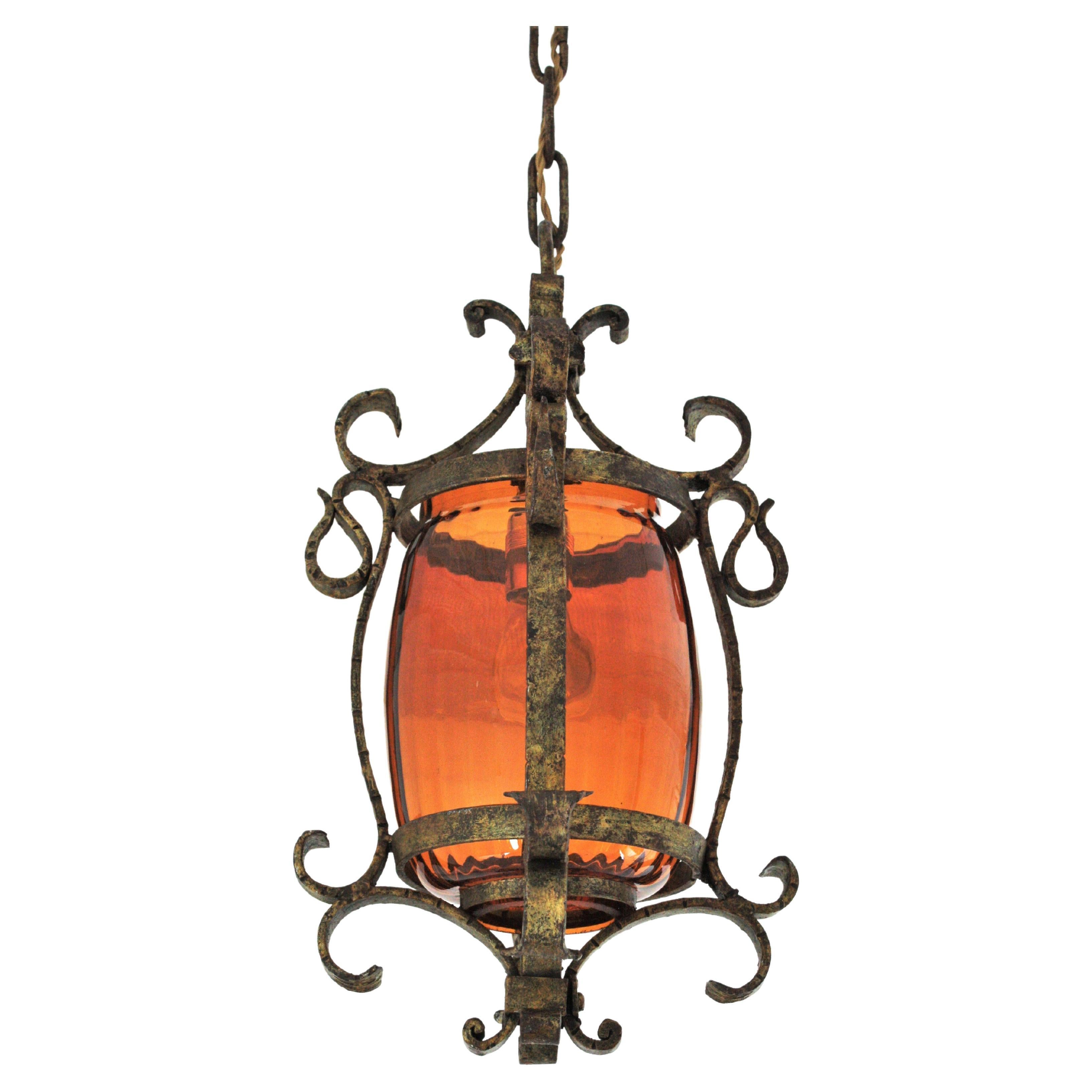 Sculptural gilt iron pendant / lantern with amber blown glass cylinder lampshade, Spain, 1930s.
This spanish hanging light features a wrought iron structure with scroll details hanging from a chain topped by a git iron canopy. An inner blown glass