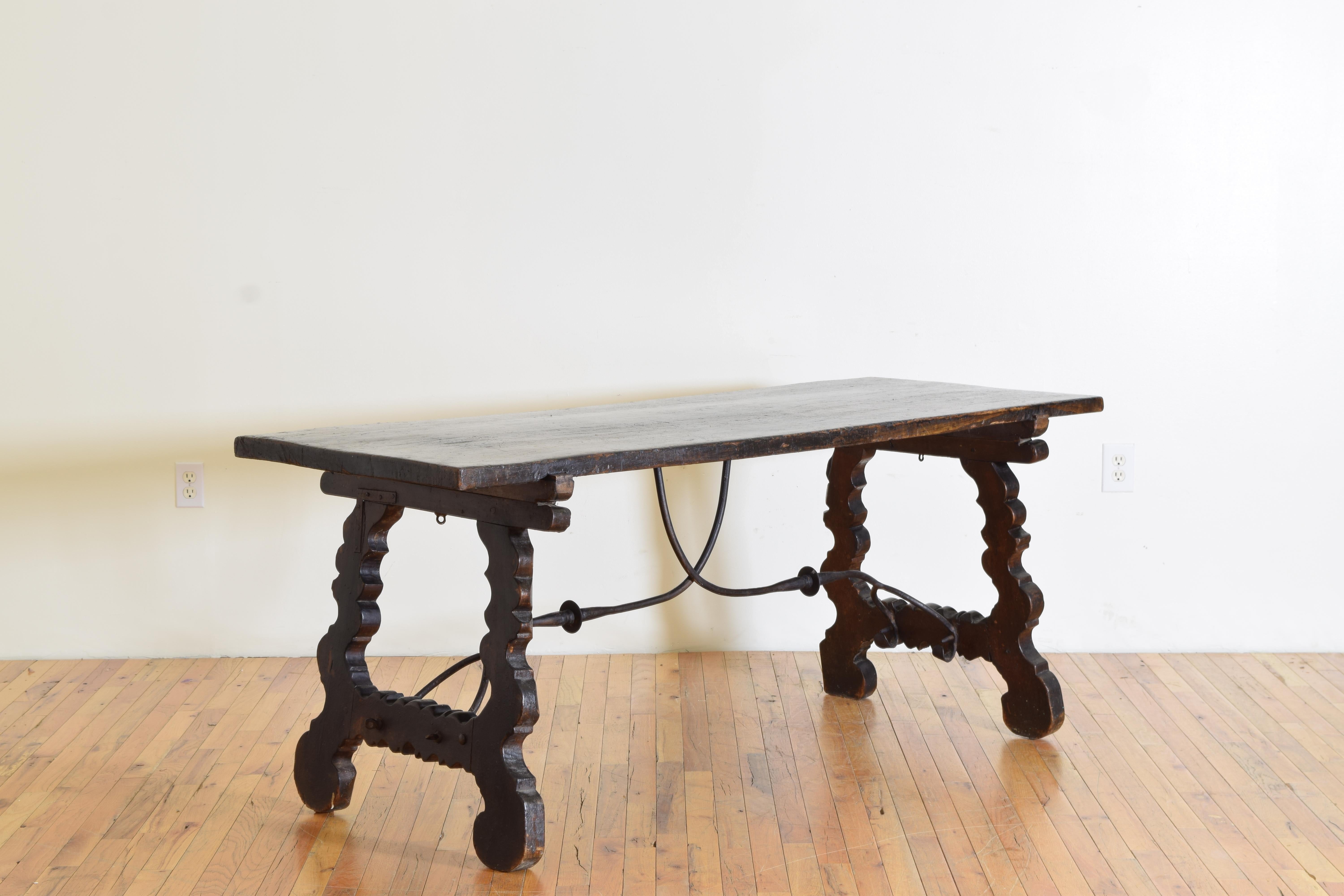 constructed entirely of dark walnut and iron, having a solid one board top supported by carved trestle-form legs joined by likely its original double pronged wrought iron stretcher