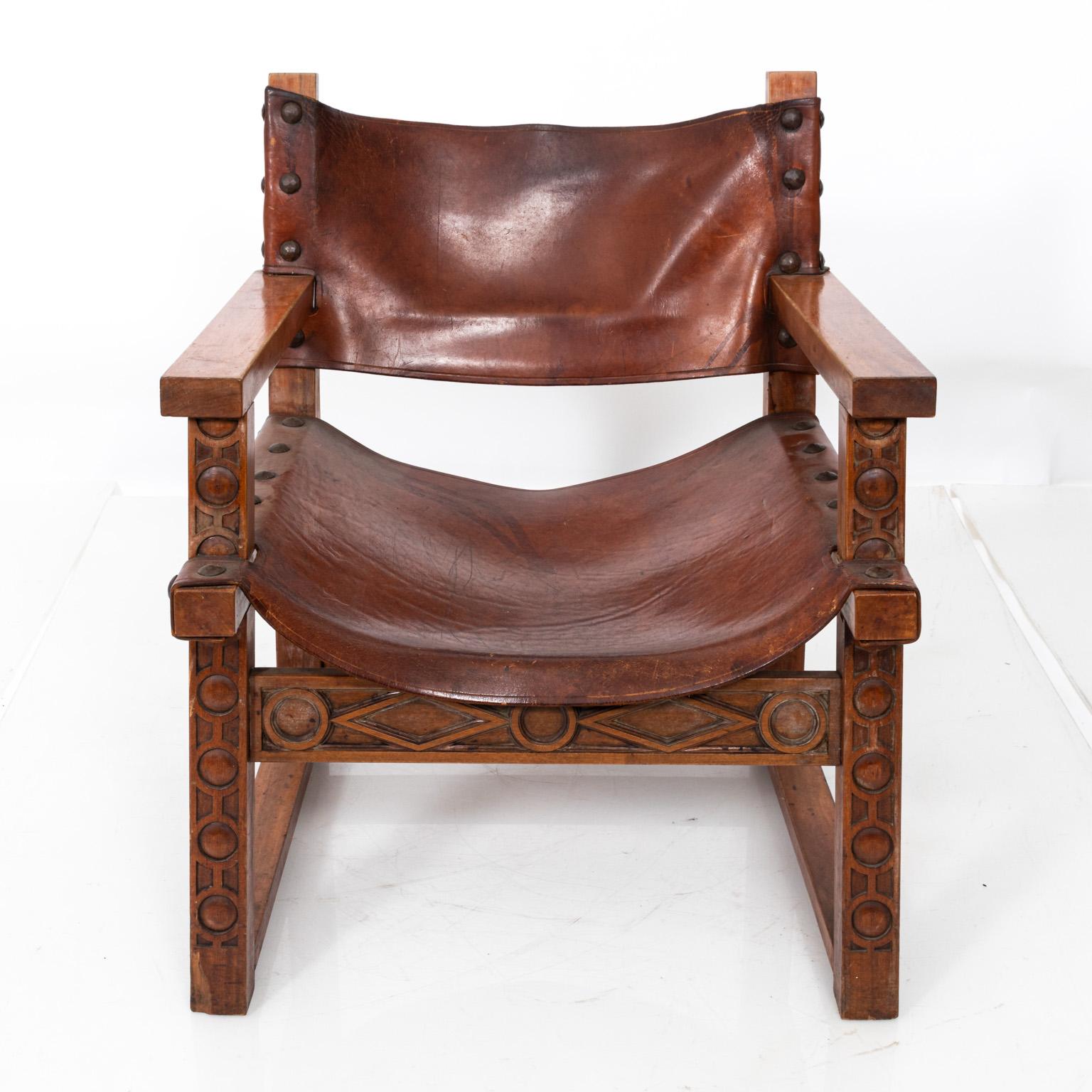 Spanish leather armchair with metal nailhead trim and carved wood frame featuring geometric shapes. Please note of wear consistent with age including some discoloration and minor tears in the leather.