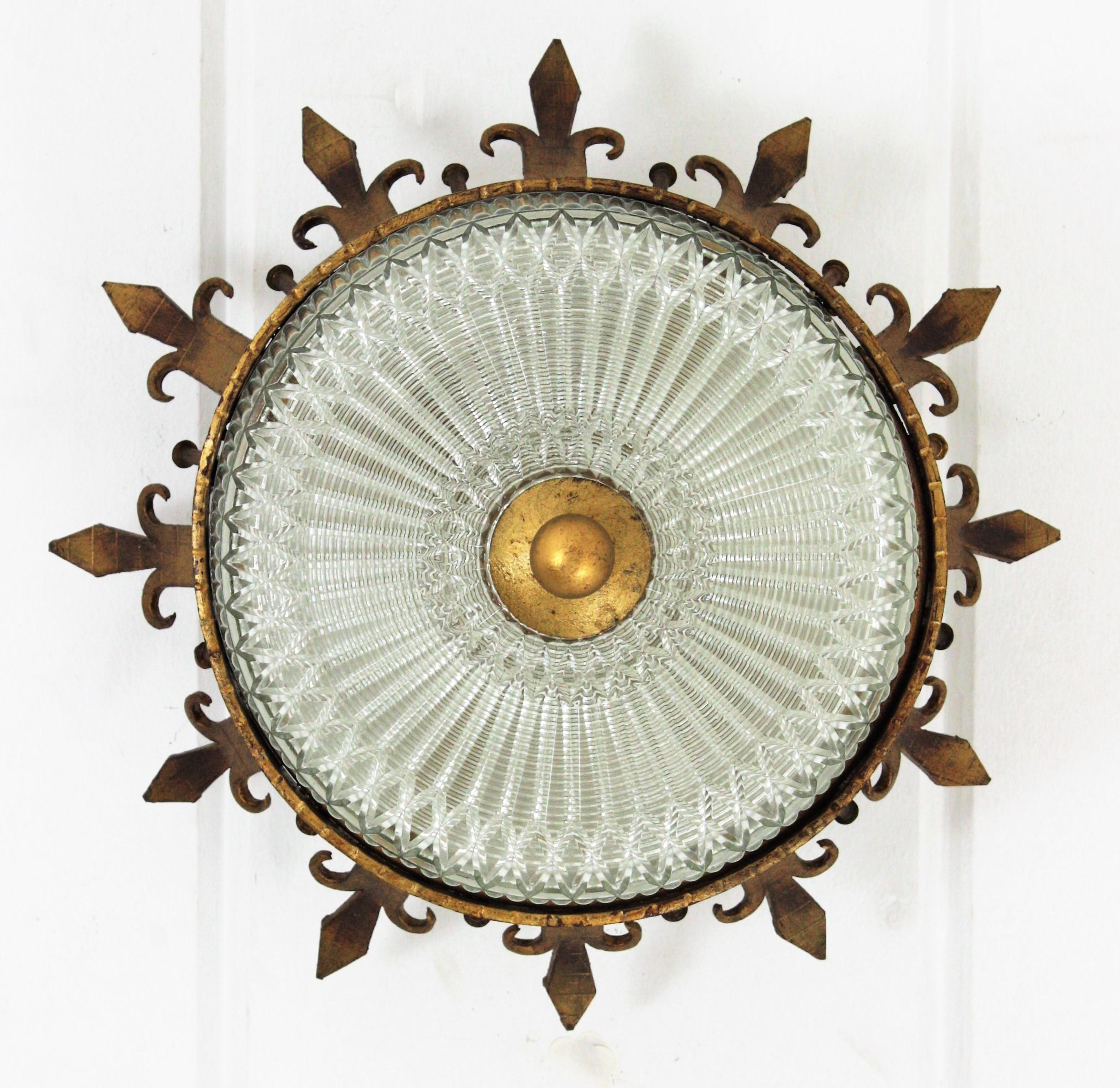 An sculptural hand hammered iron and glass ceiling light fixture with neoclassical and gothic revival accents. Spain, 1930-1940s.
This hand forged and gilded iron crown sunburst light fixture has a fluted glass shade with a gilt iron ball finial.