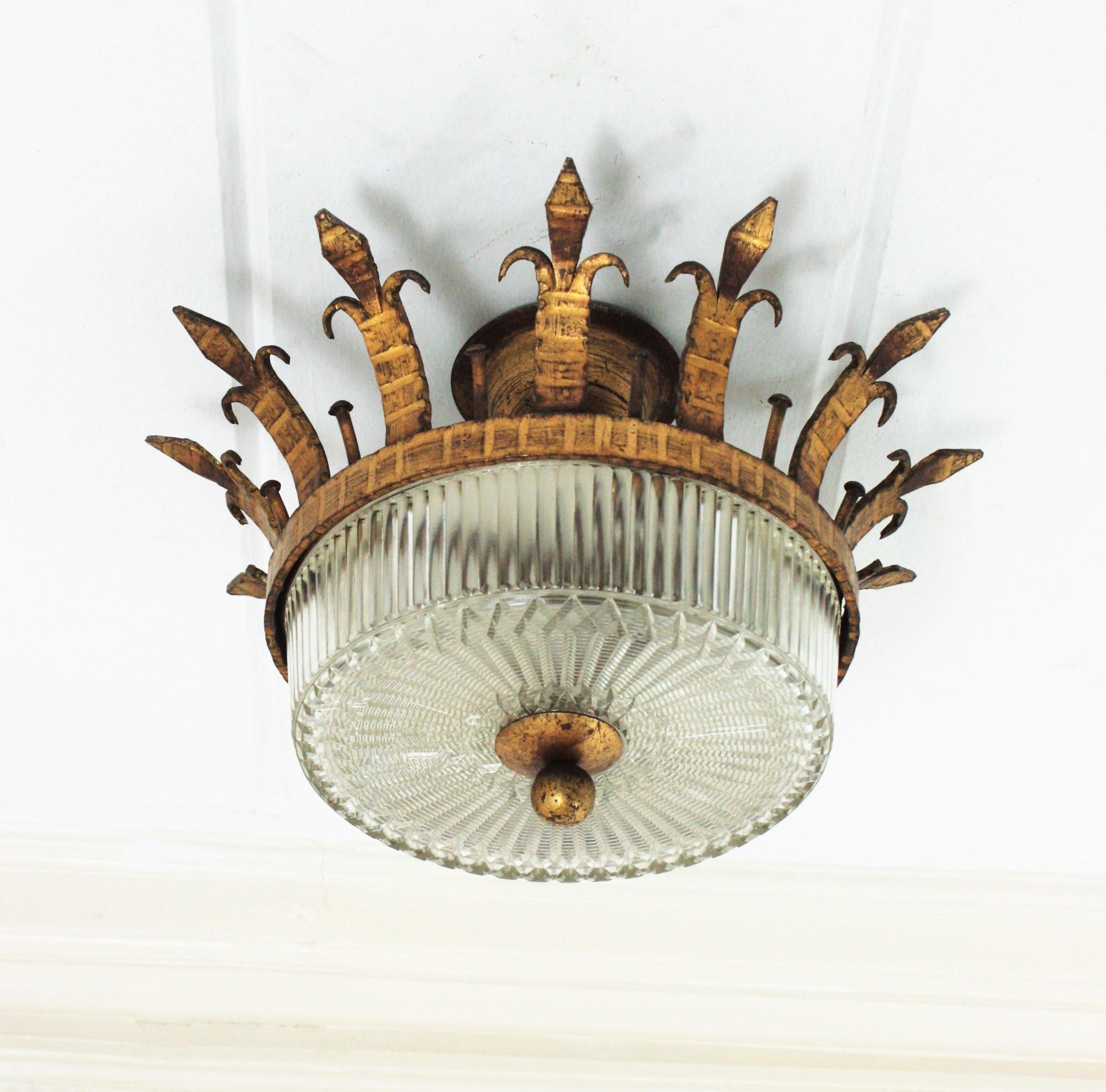 Monture à couronne en fer forgé et verre pressé, Espagne, années 1930-1940.
Un plafonnier sculptural en fer martelé à la main et en verre aux accents néoclassiques et gothiques. 
Ce luminaire en forme de couronne en fer forgé et doré est doté d'un