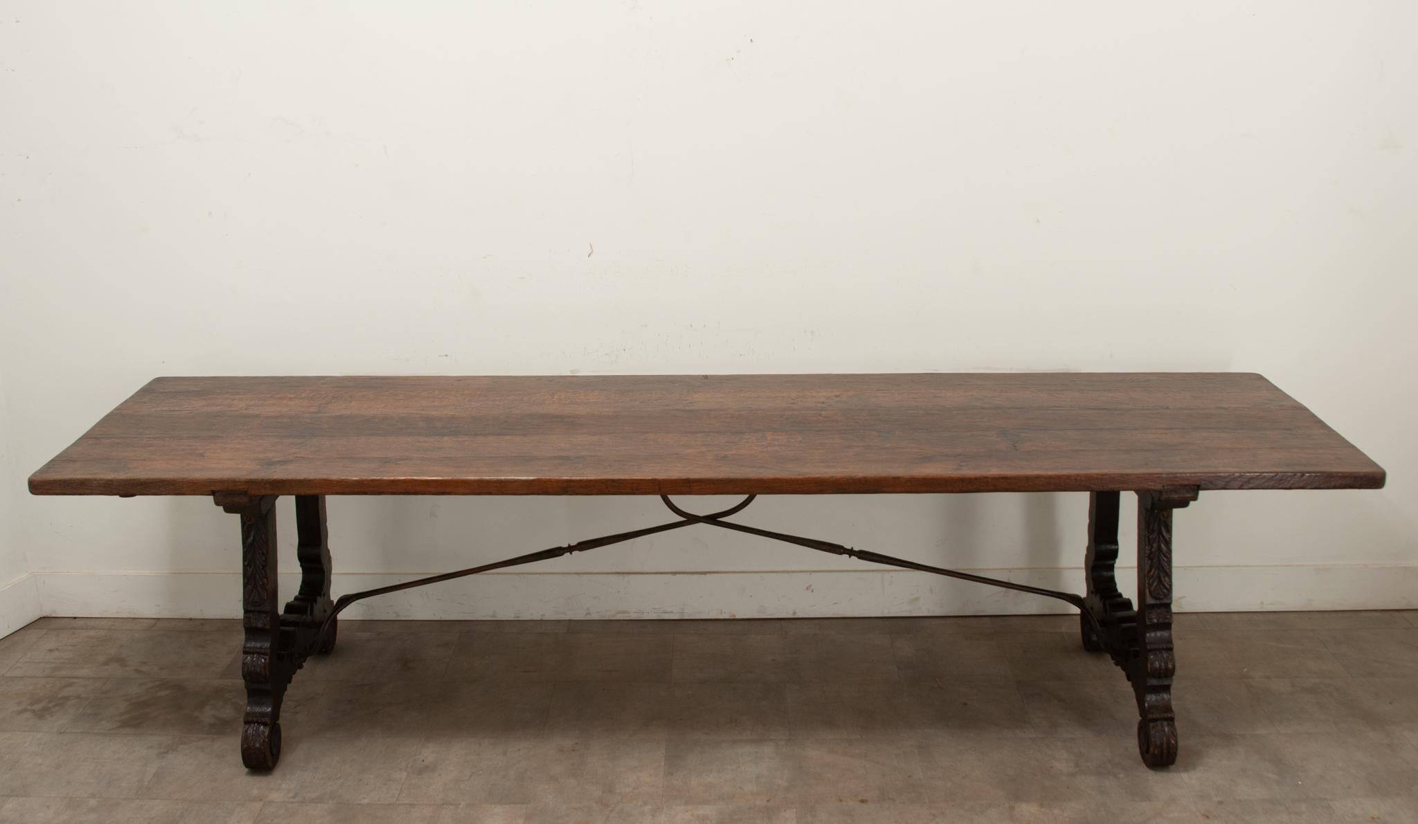 A large oak Spanish dining table extends to over twelve feet long. This table has a simple plank top over carved decorative legs connected by a hand forged iron support. At each end you’ll find iron brackets under the table top that the removable