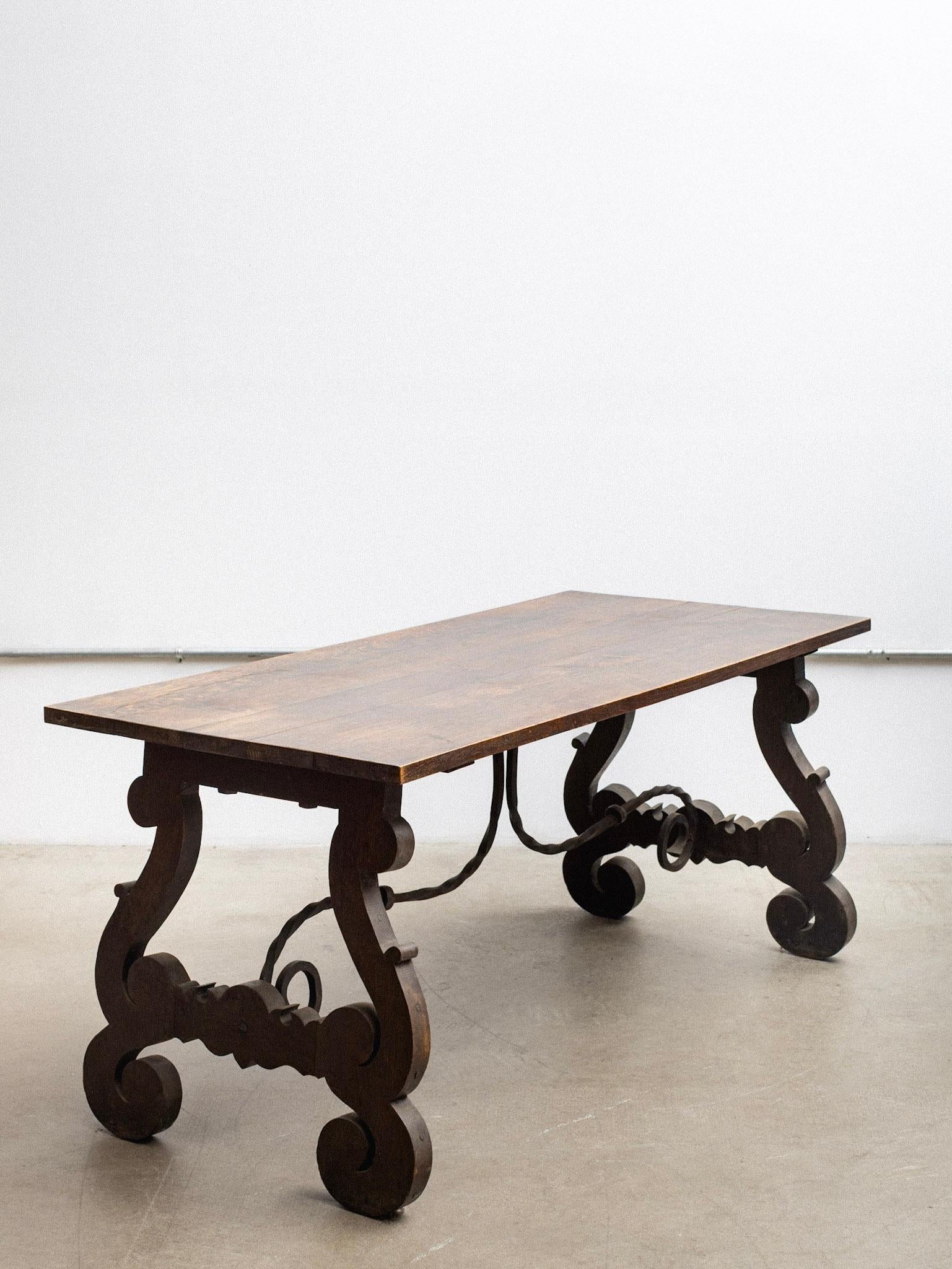 Table de salle à manger en chêne espagnol avec un plateau rectangulaire et une base à tréteaux à volutes. Les pieds sculptés sont reliés à des châssis en fer forgé. La finition teintée foncée met en valeur les veines naturelles du bois.