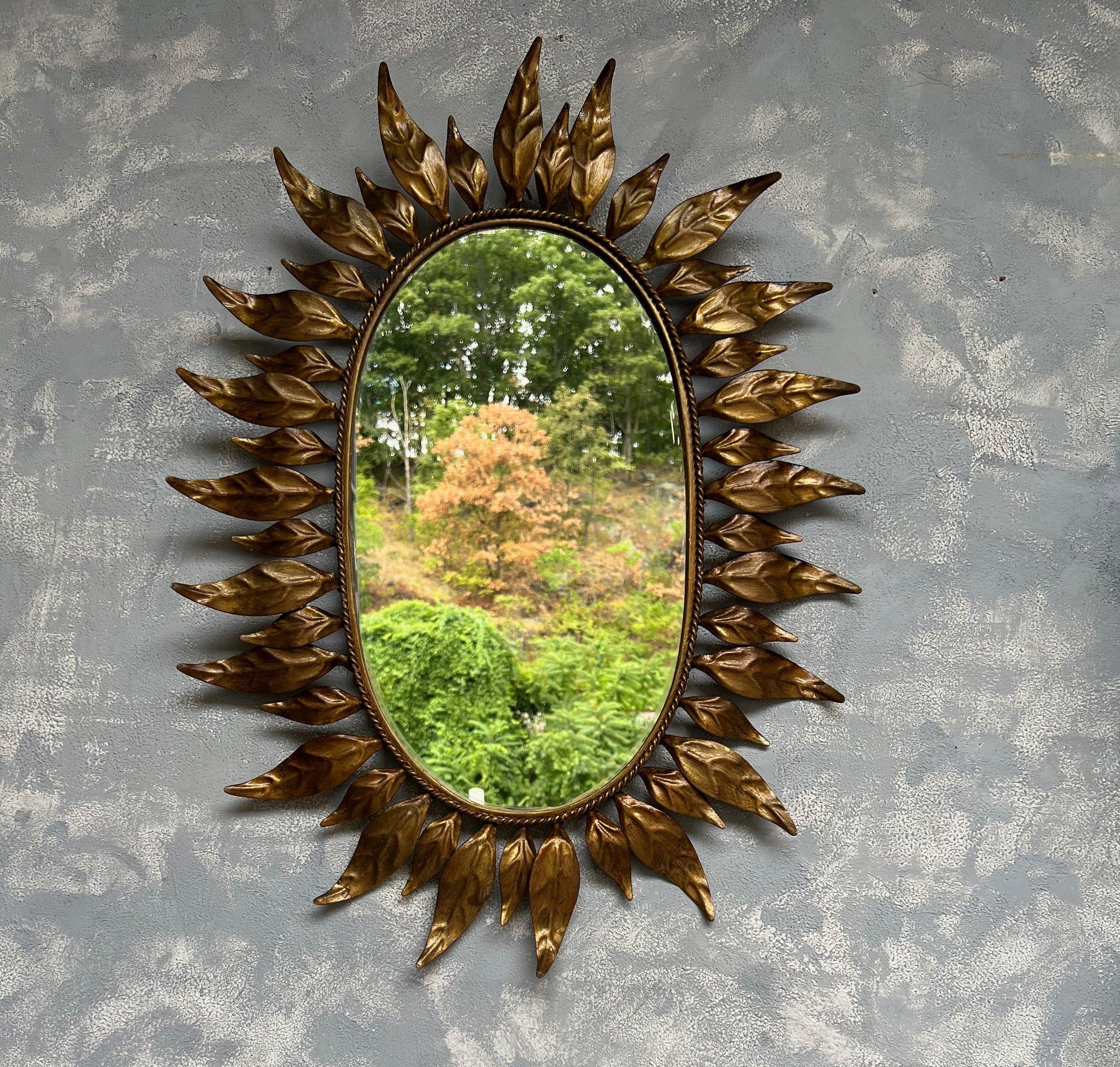 Dieser ovale Spiegel aus vergoldetem Metall besticht durch sein ausgeprägtes und fesselndes Design. Der Spiegel zeigt eine Kombination aus großen und kleinen Blättern oder Strahlen, die einen fein geflochtenen Innenrahmen umschließen. Das Metall