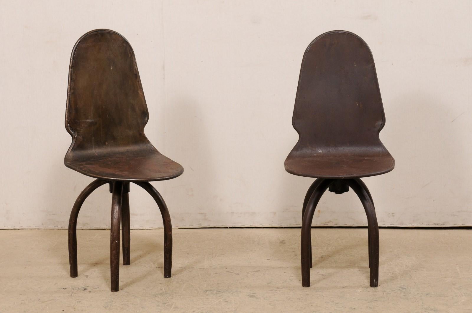 A Spanish pair of iron swivel chairs. This vintage pair of chairs from Spain are fun and quirky! They are constructed from iron and feature arched backsides with notched accents at the hips. The backs roll as one continuous piece onto the seat below