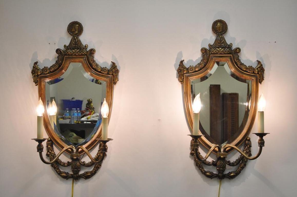Pair of stunning Spanish Renaissance style cast brass, wood, and bevelled mirror electrified wall sconces. Item features shield form solid wood frames, cast brass floral, acorn, and cameo accents, bevelled glass mirrors, two electrified arms each.
