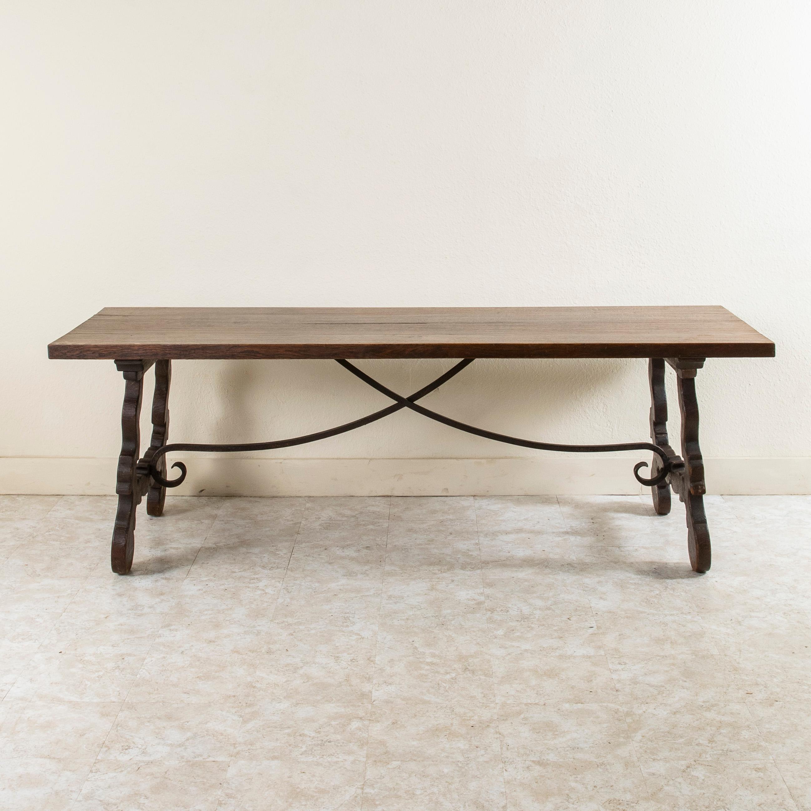 This large Spanish Renaissance style oak dining table from the turn of the twentieth century features a hand forged iron trestle and a hand-hewn 1.75 inch thick top constructed of four planks of wood. Its thick solid oak construction and dark patina