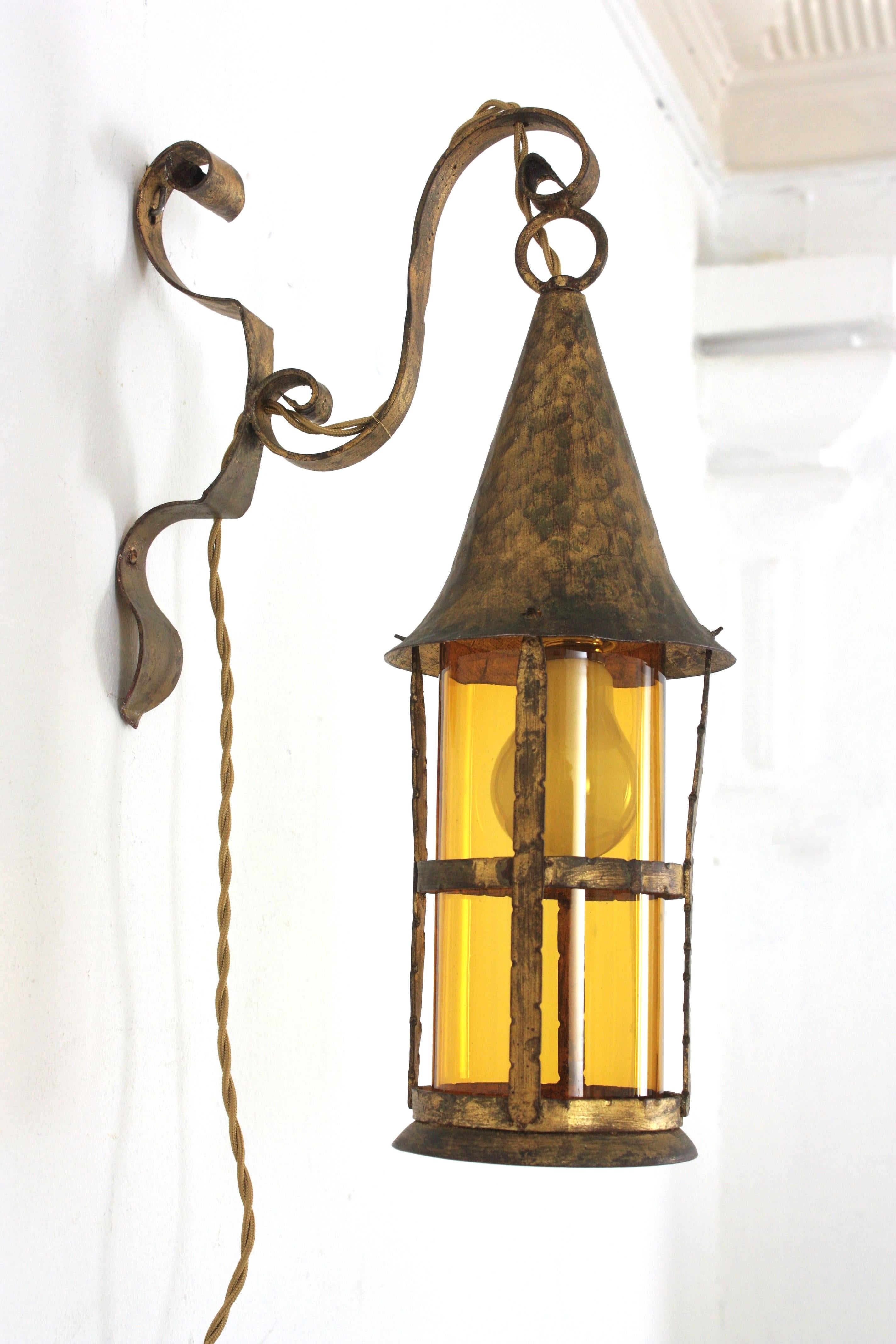 Spanish Revival Hand Forged Wall Hanging Lantern / Wall Sconce in Gilt Iron
Sculptural gilt iron wall sconce / lantern with amber blown glass cylinder lampshade, Spain, 1940s
This spanish hanging light features a wrought iron lantern with conical
