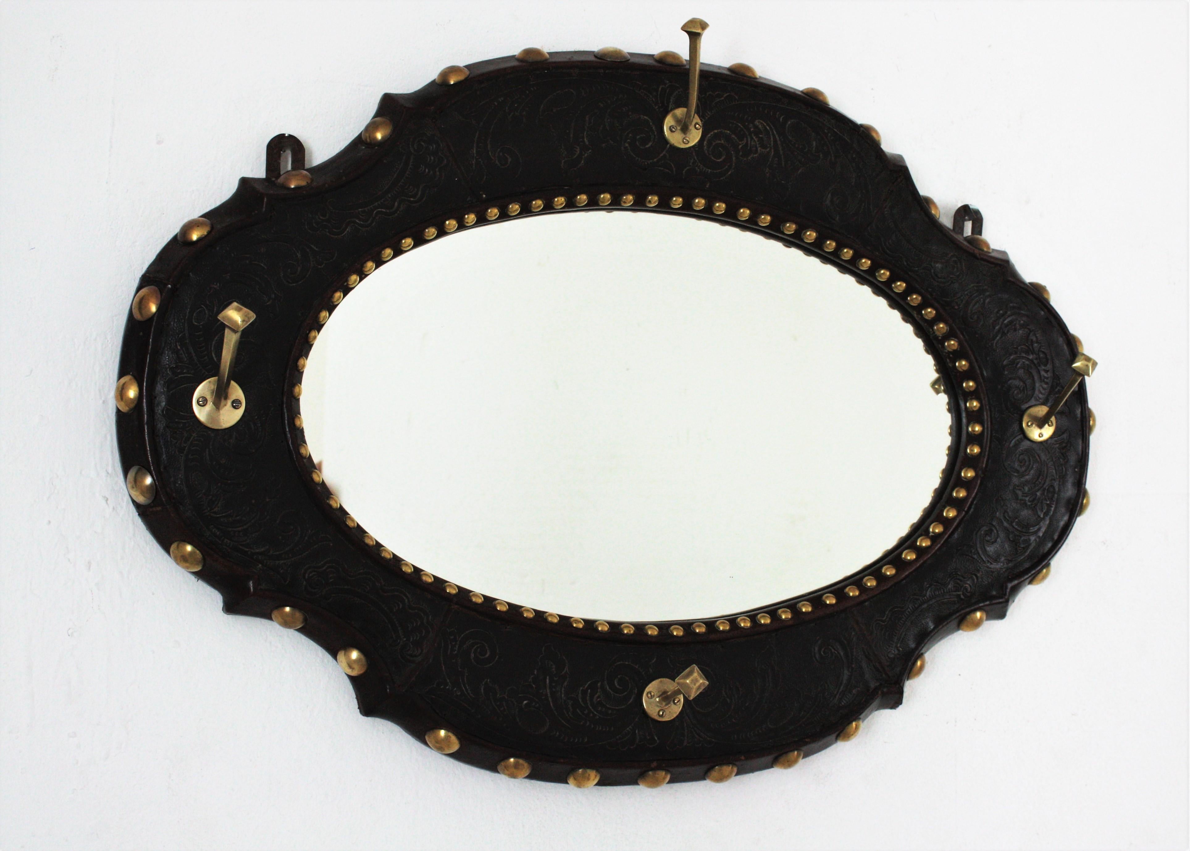 Elegant Spanish Colonial style wall mirror with coat hangers in leather repoussé and brass. Spain, 1940s.
This piece features an oval beveled mirror with a leather repoussé frame with foliage decorations and 4 brass coat 
hangers. 
Nicely