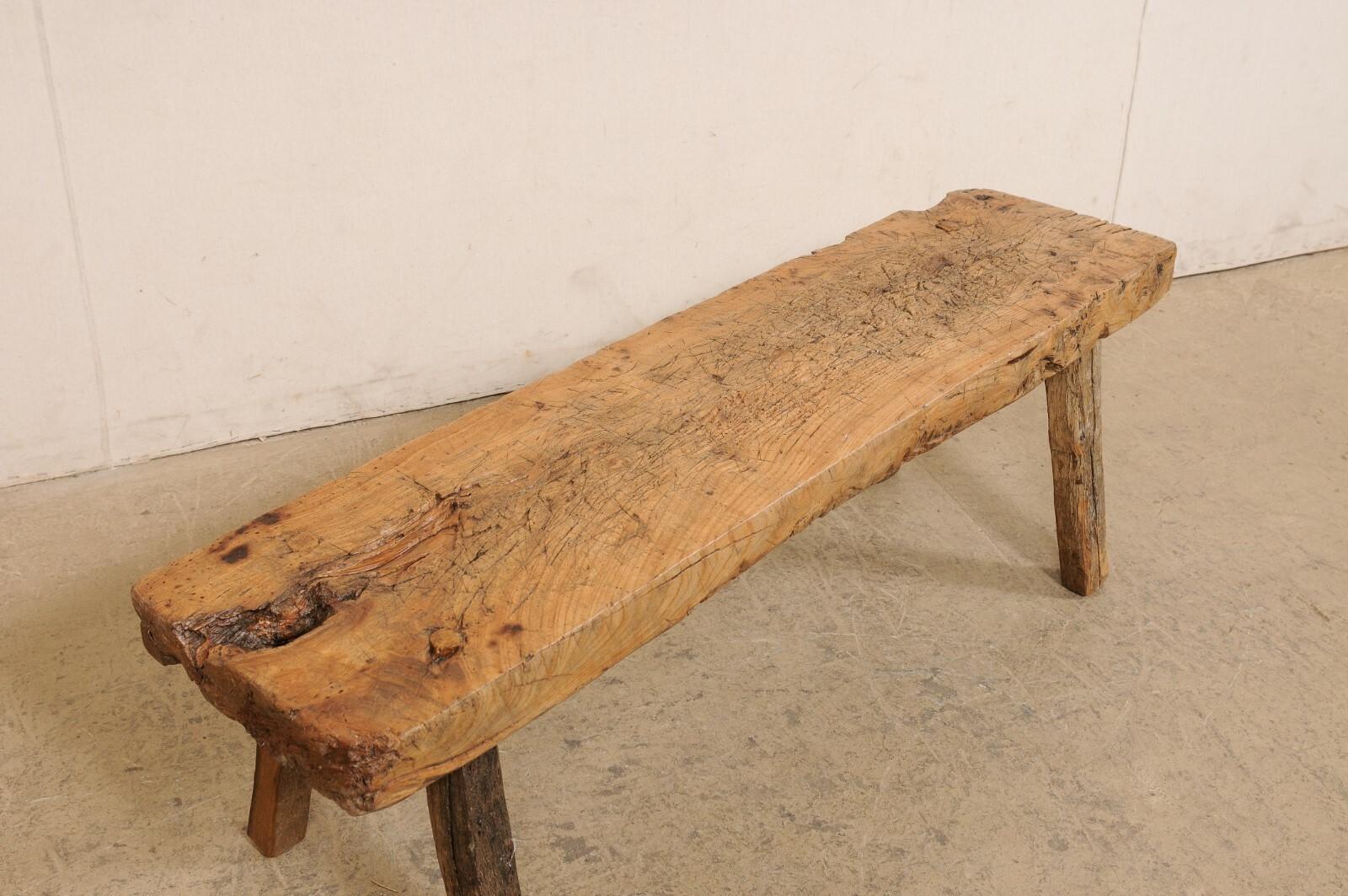 A Spanish rustic wooden bench. This vintage bench from Spain, just over 4.5 feet in length, has been constructed of a single and thickly cut slab board at top/seat, rectangular in shape, and supported by four squared legs that cant outwardly. The