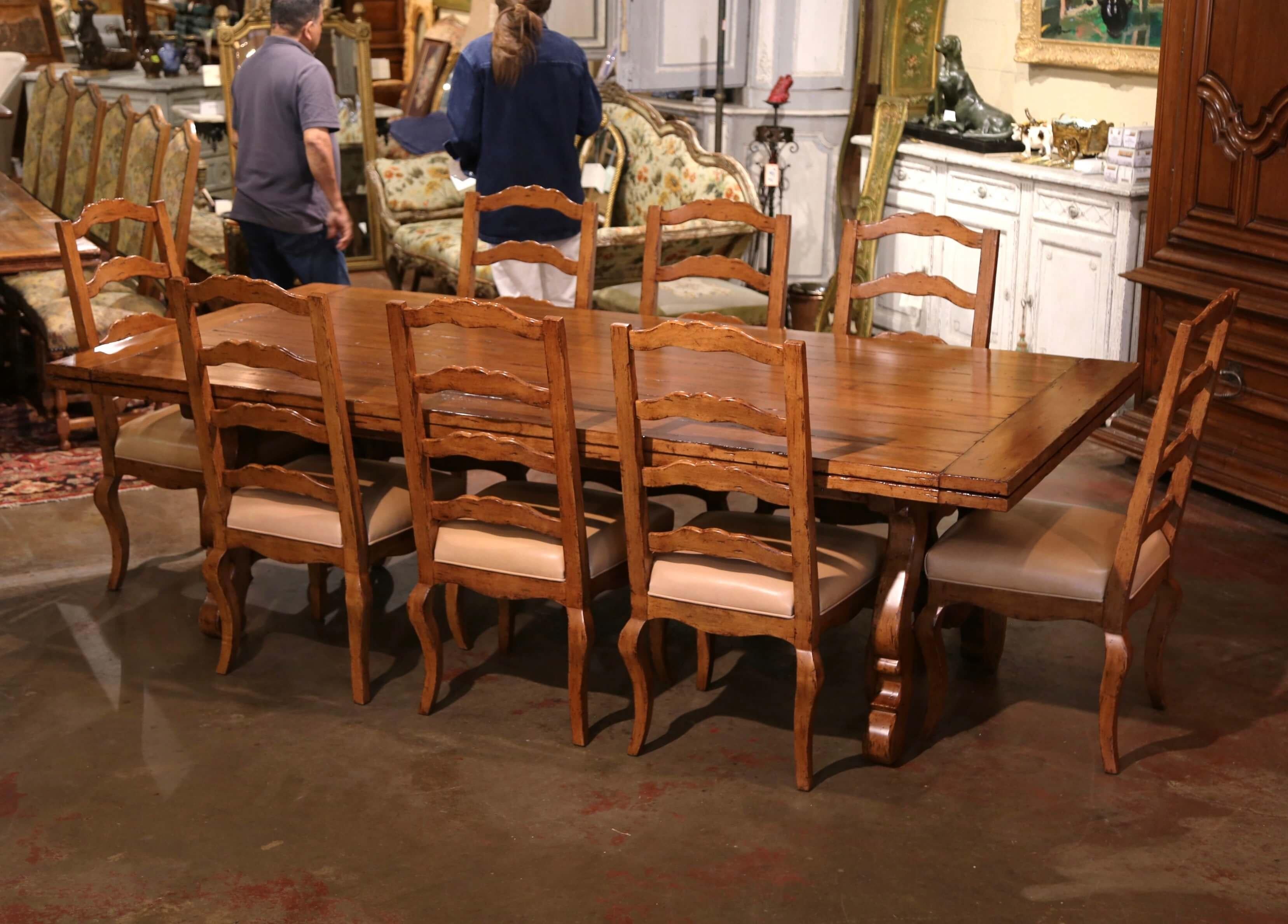 A fabulous dining room set comprising of a large and wide table with leaves and matching 12 ladder back chairs with leather seat. Standing on two intricate scrolled pedestal legs connected with a carved stretcher, the rectangular top is built with