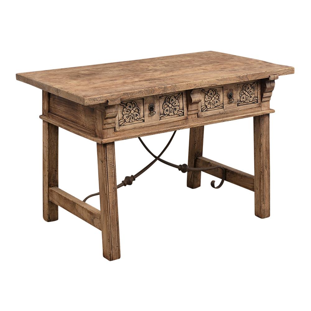 This Spanish Colonial style library table features a bleached wood finish a thick solid wooden top. The table has two drawers with carved butterfly designs, and stretched legs that are joined by decorative wrought iron crossbars. This table is