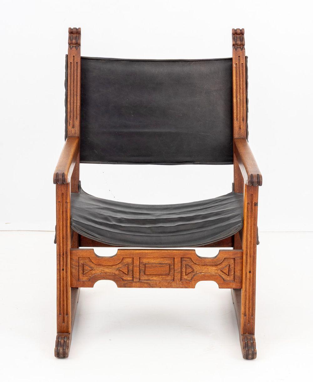 Spanish Style Walnut Armchair, with black leather and riveted seat and back rest, with geometric carved apron on four carved wood legs conjoined by stretchers. 39.25