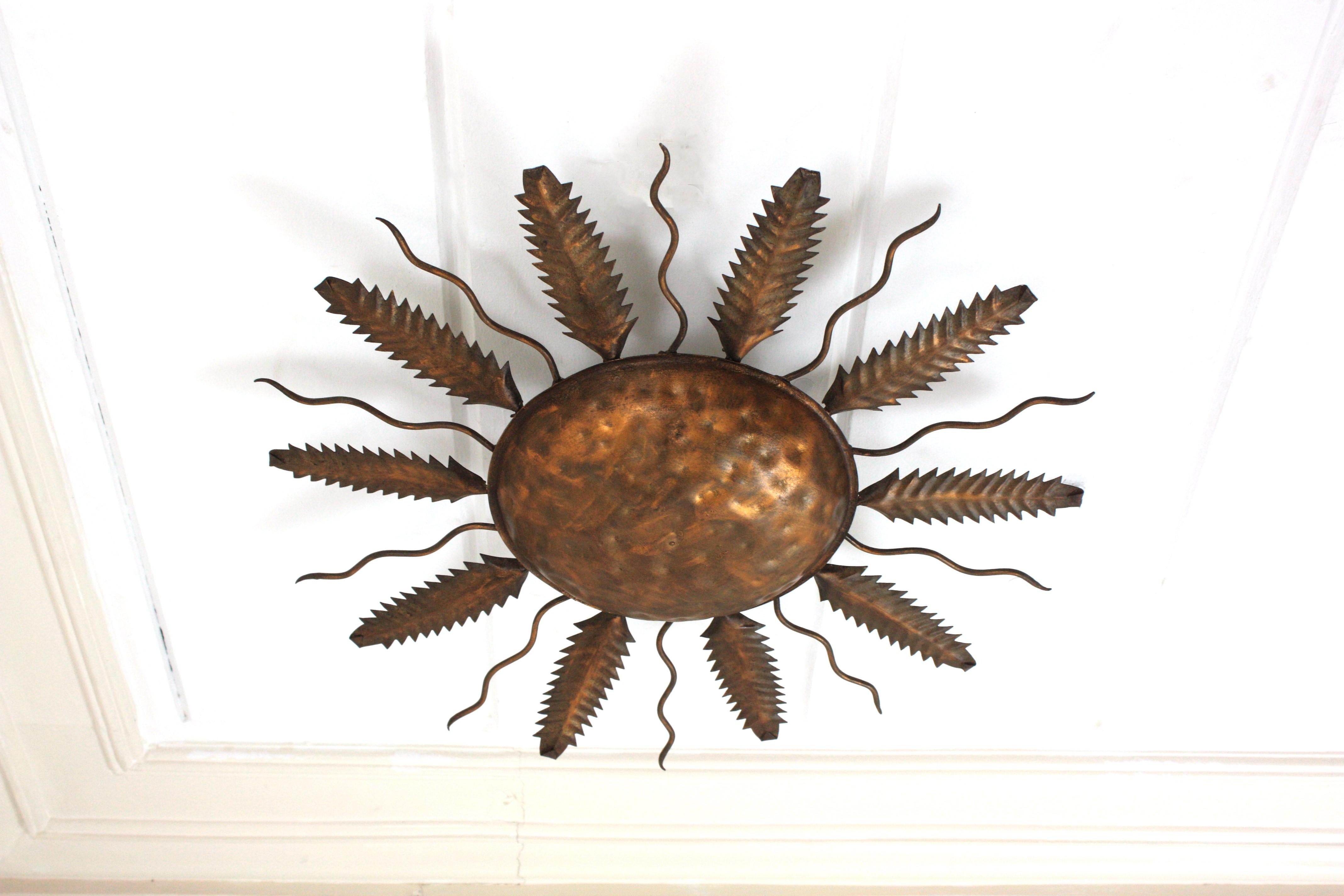 Large gilt metal leafed sunburst flush mount. Spain, 1950-1960.
This stunning ceiling flush mount light fixture features an iron structure with a central round panel surrounded by alternating jagged leaves and curly rays.
The central panel is richly