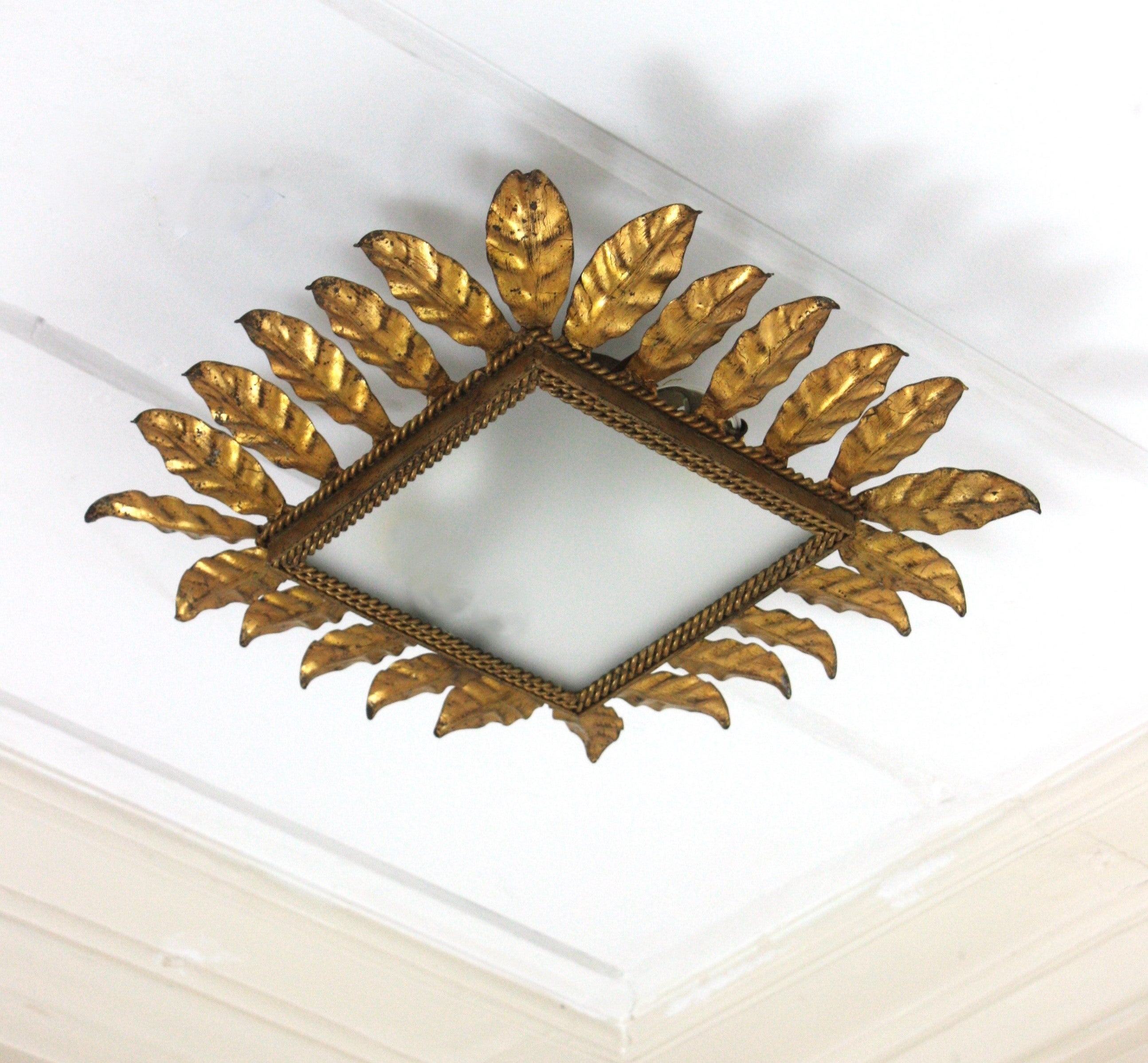 Sunburst leafed  flush mount with gold leaf gilding and frosted glass shade. Spain, 1950s.
Square shaped foliage panel supporting the frosted glass shade. The central glass is surrounded by a double twisting iron rope and layer of hand hammered