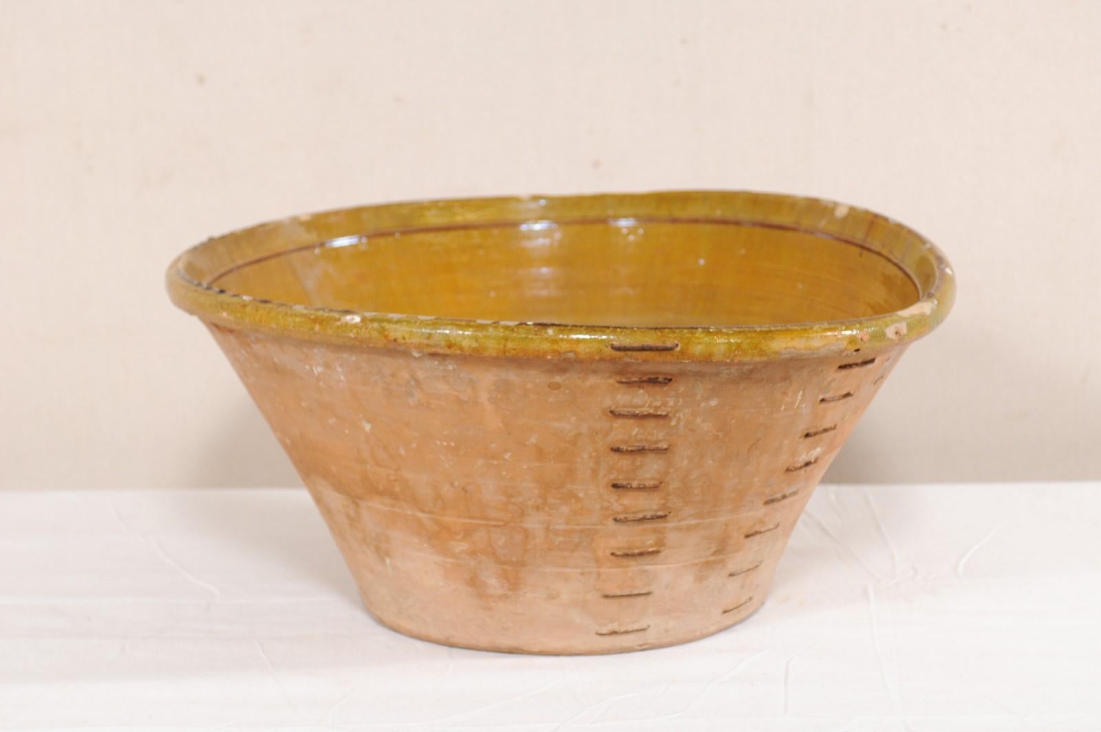 A Spanish terracotta bowl with muted yellow and pale green hues from the early to mid-20th century. This bowl from Spain is round in shape, with widest part being at the top lip, it gradually tapers towards the bottom, and rests upon a flat base.