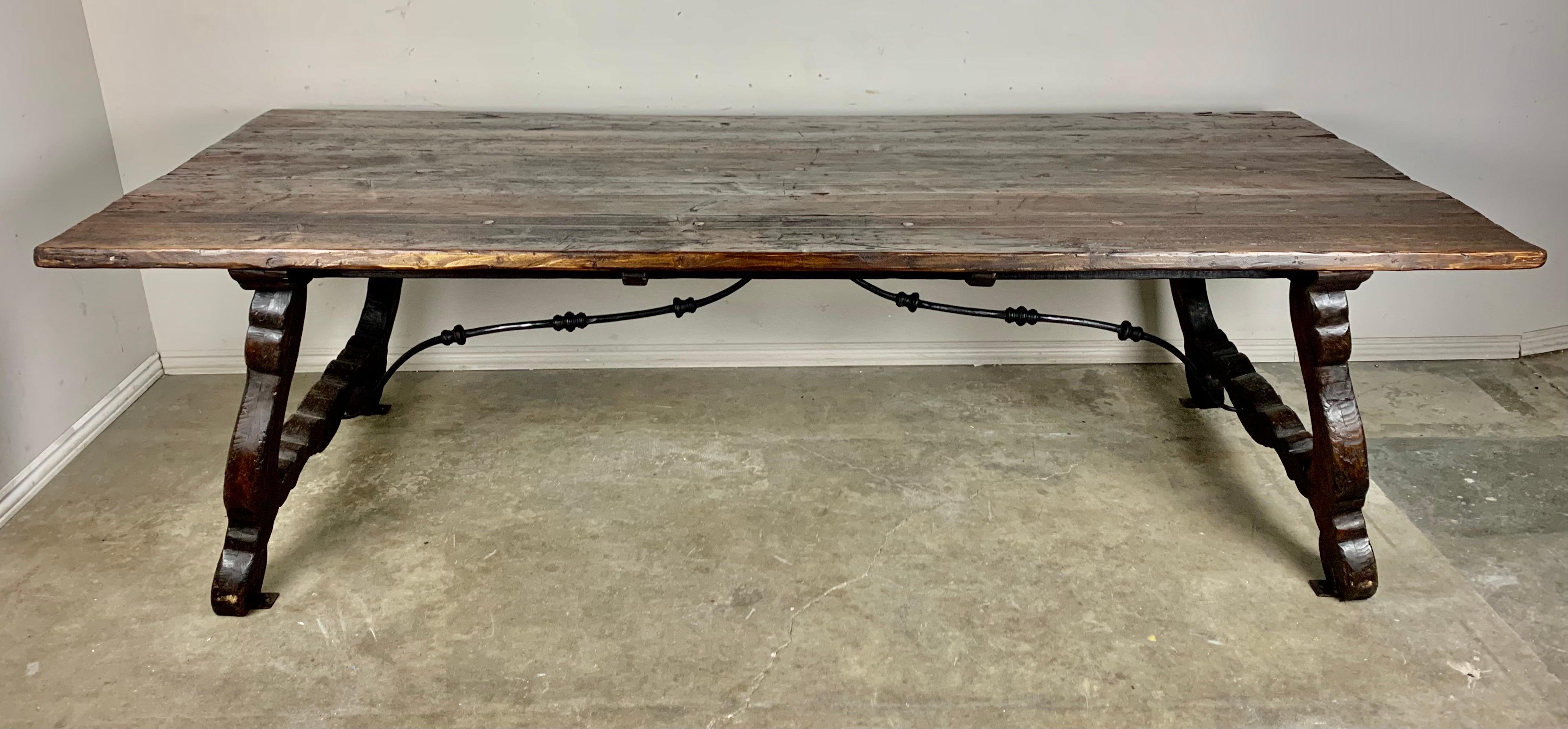 Spanish trestle style dining table standing on two lyre shaped pedestals. Handwrought iron stretcher connects the lyre pedestals. The large rectangular shaped top shows distressing including cracks and repairs, but I think it adds character to a