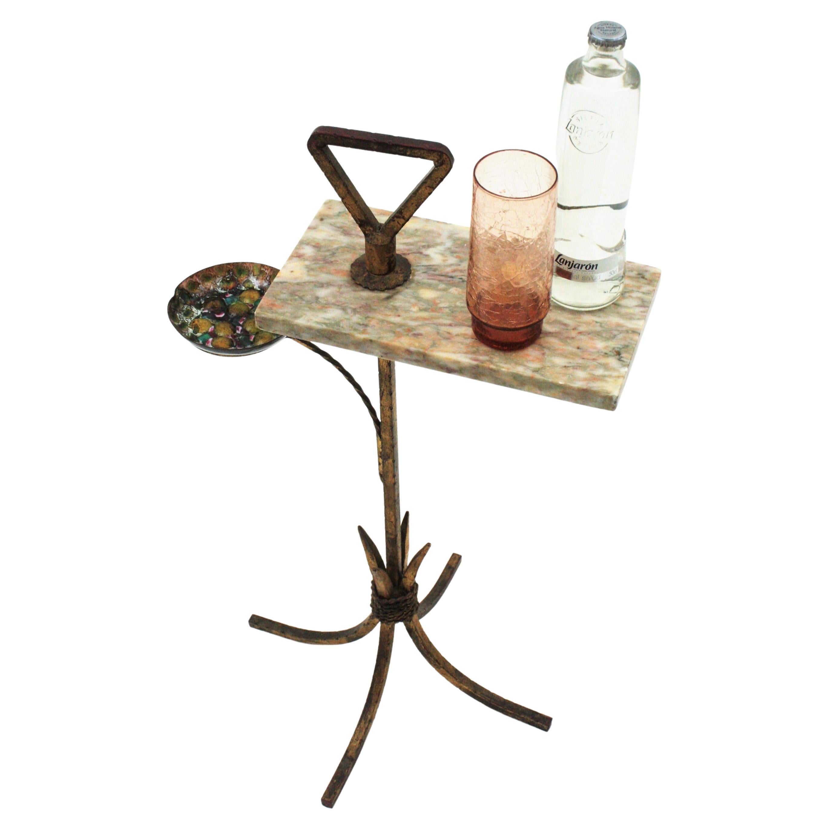 One of a kind gilt iron and marble end table with colorful enamel ashtray. Spain, 1940s-1950s.
This stylish hand forged iron table features a grey and white marble tabletop standing on a git iron structure with a four footed base. The marble top