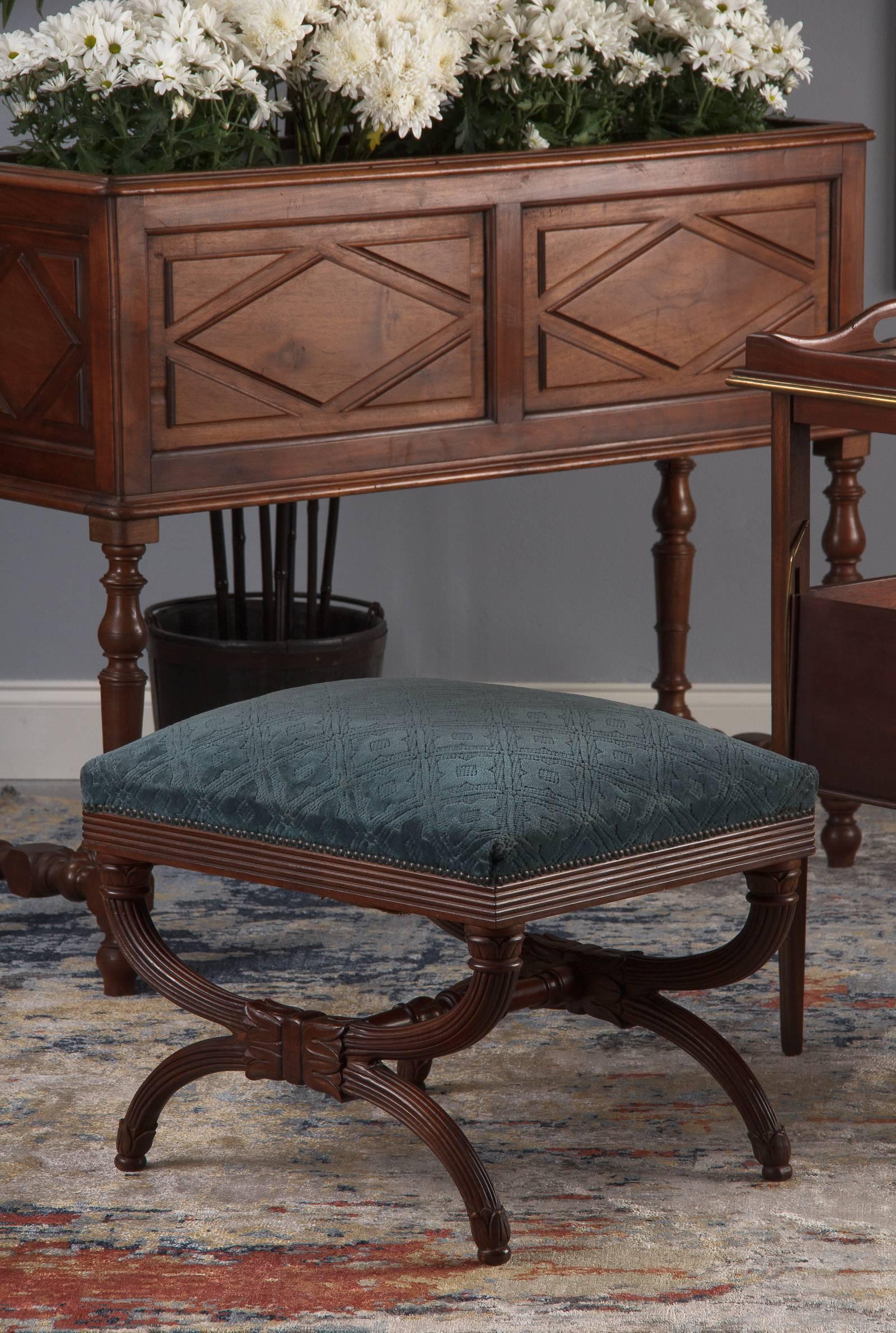 An ornate upholstered Spanish savonarola ottoman in carved walnut, circa 1920. Walnut base with original velvet upholstery over spring seat. The base has a savonarola X-form shape with carved ribbing around the bottom of the seat and along all the