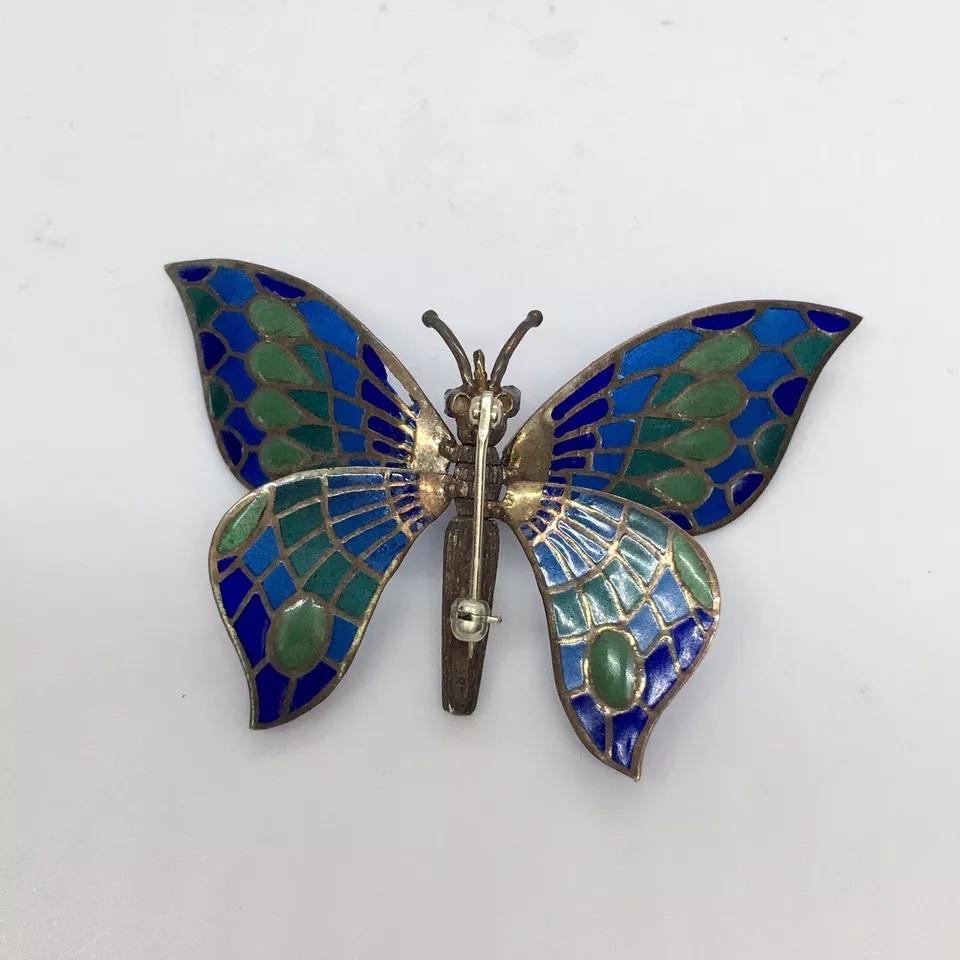 This vintage 18k white gold brooch features a beautiful butterfly shape with multicolor sapphire stones and a 0.06 carat ruby content. The enamel work is exquisite, showcasing a plique-a-jour style that adds depth and dimension to the piece. The