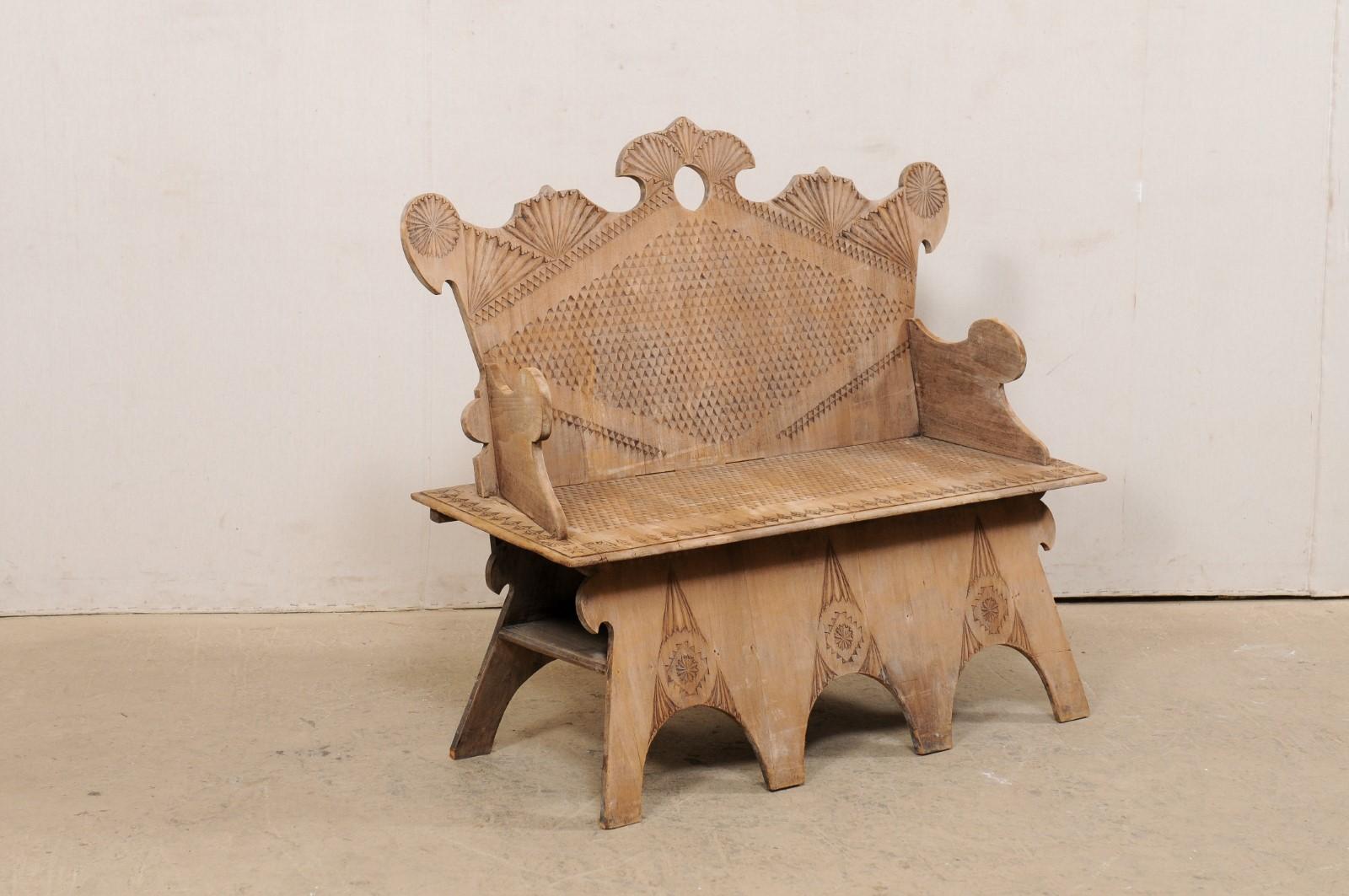A Spanish carved wooden arm-bench with back from the mid 20th century. This vintage natural wood bench from Spain is adorn with Moorish influenced carvings, featuring a high back with undulating crest in a fan-carved motif with pierced center. The