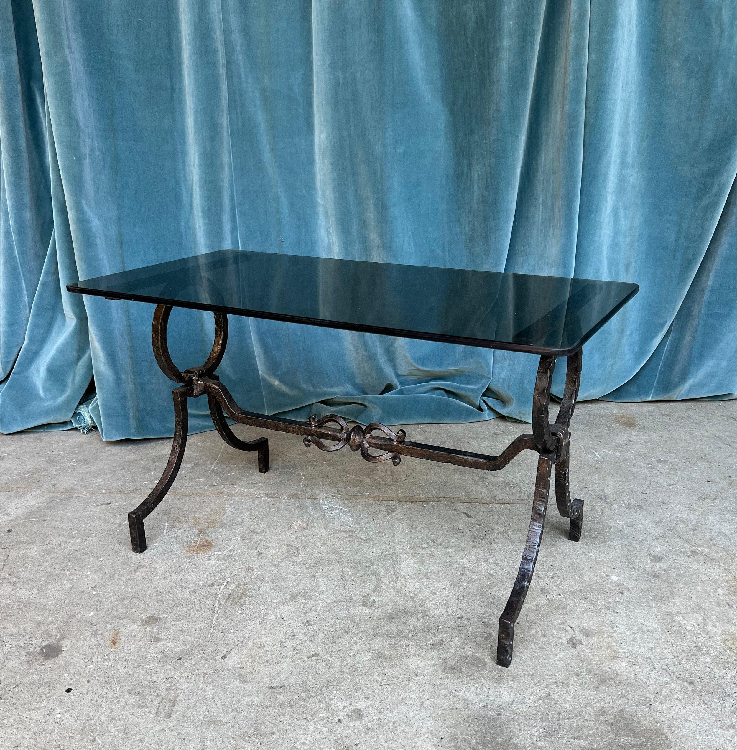 This striking 1950s Spanish wrought iron coffee table with smoked grey glass exudes timeless elegance and sophistication. The handsome black iron base, adorned with large circular elements and elevated on gracefully curved legs, perfectly