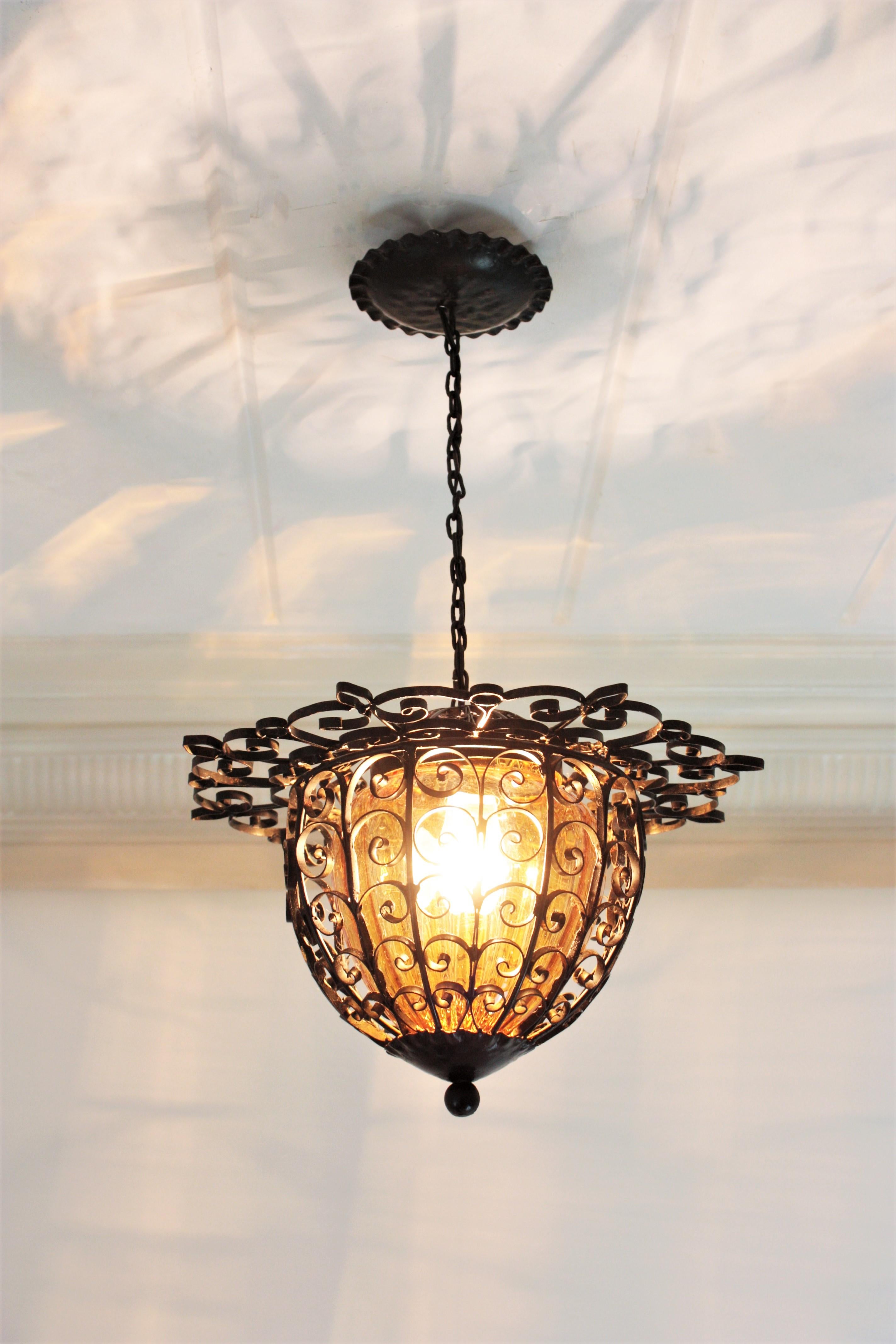 Eye-catching scrollwork iron lantern or hanging light with hand blown amber shade, Spain, 1940s.
The structure of this pendant light is made with wrought iron scroll decorations. It has an inner amber glass globe shade and the top of the lantern