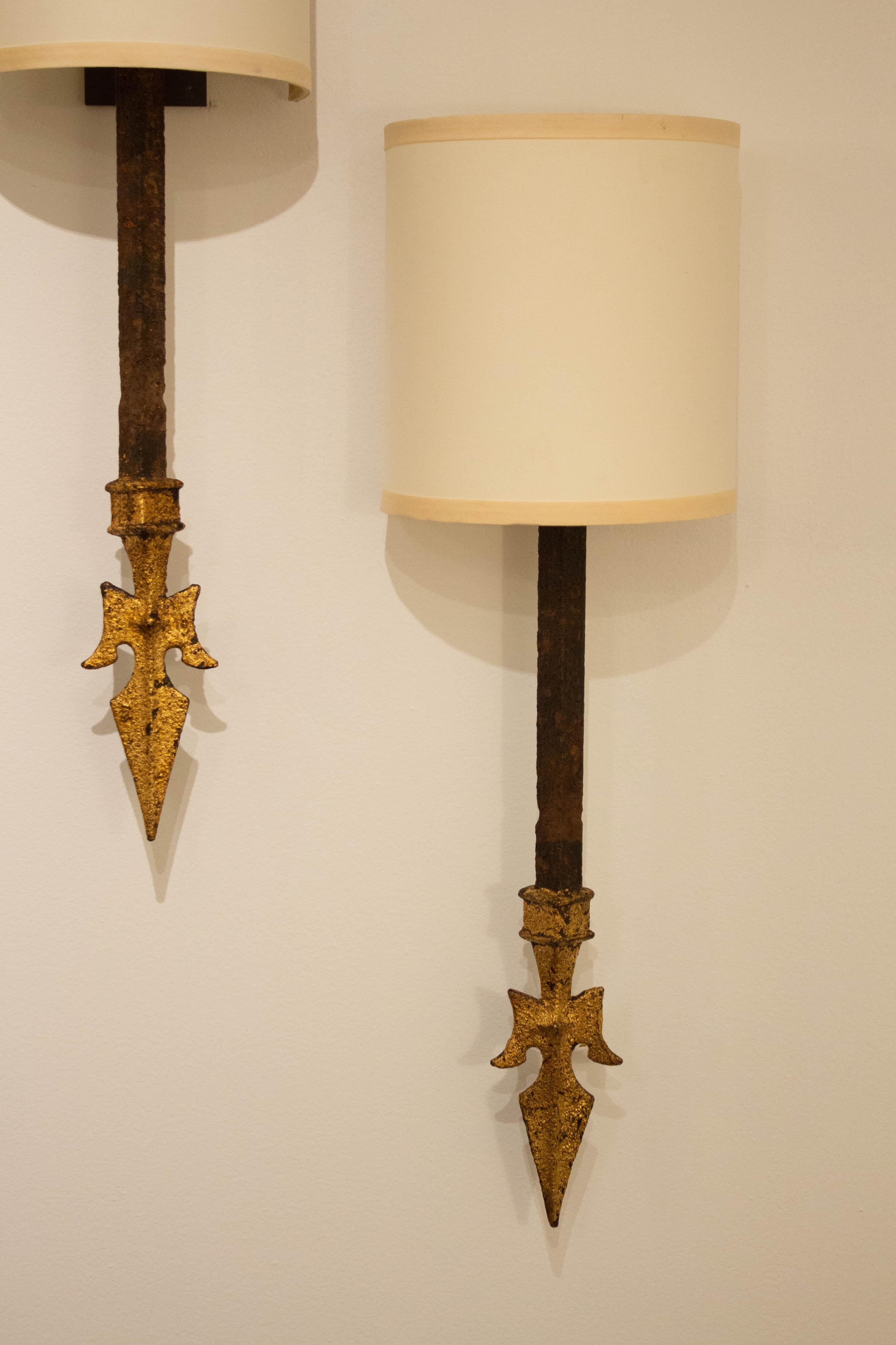 A pair of wrought iron wall sconces. Gold gilt spear point finials. Hardwired. Cream half shades included.