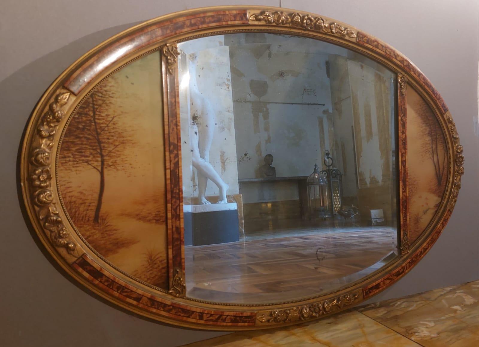 Specchiera ovale con specchio originale al centro e bordo smussato. La specchiera è divisa in tre parti: i laterali sono due vetri dipinti con scene boschive dai colori autunnali che rappresentano un ruscello con alberi e piante acquatiche. La